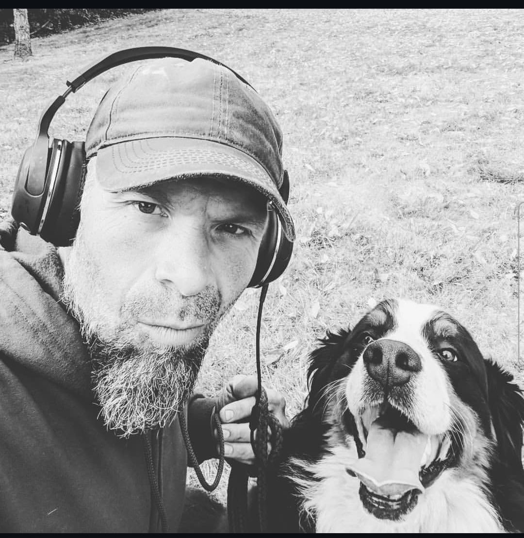 Man with headphones and beard taking a selfie with a happy dog outdoors, grassy background.