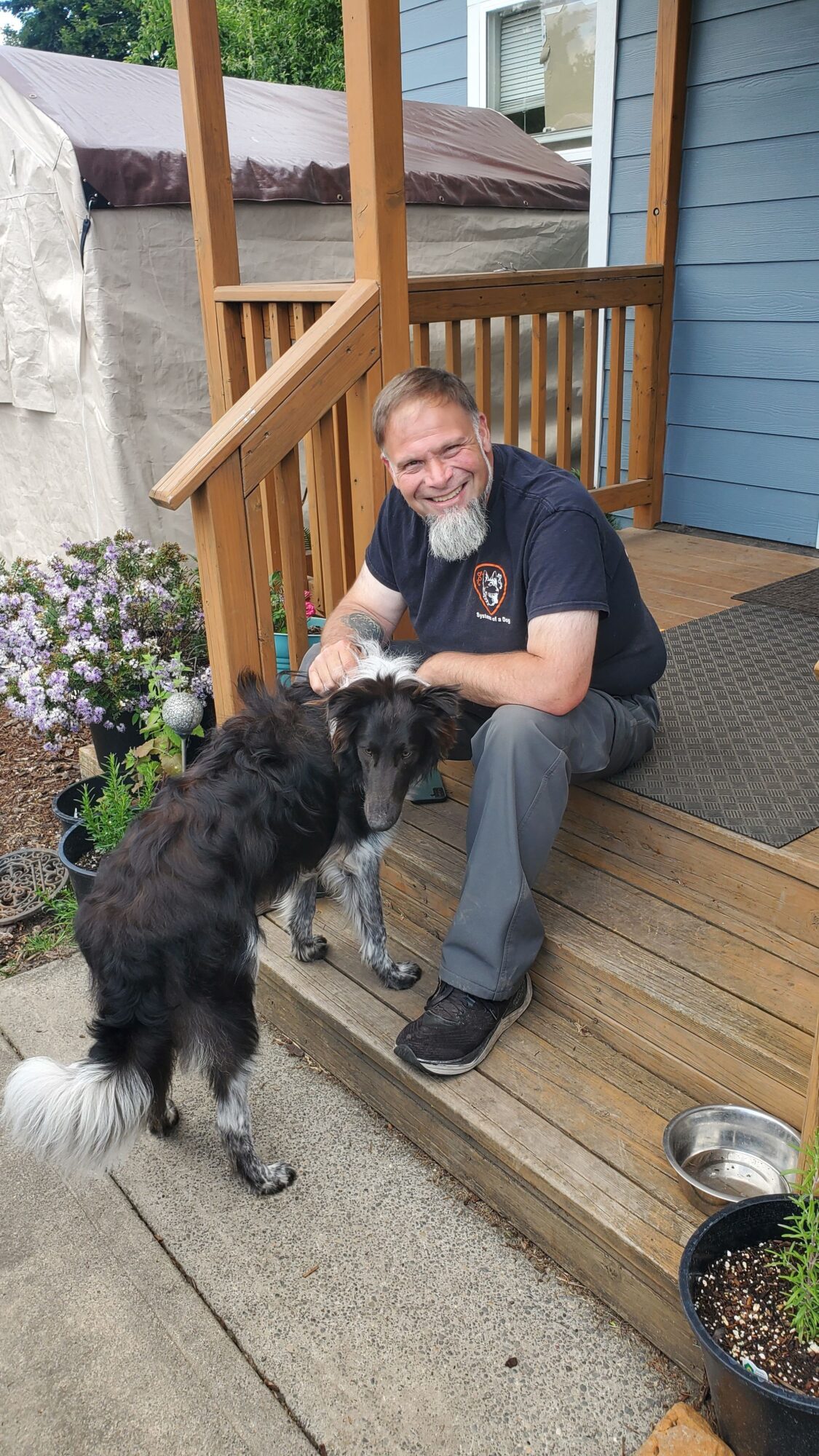Person crouching on a porch with a black and white dog, wooden railing, potted plants, and a concrete sidewalk.