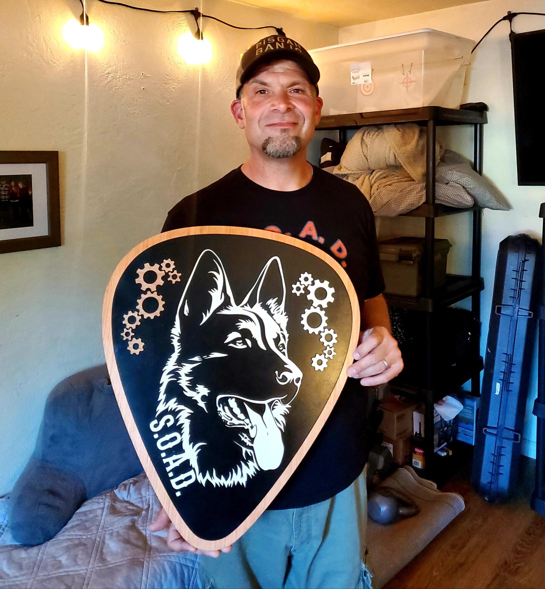 Man holding a shield with a wolf illustration and gear design, smiling indoors with shelves and a TV in background.