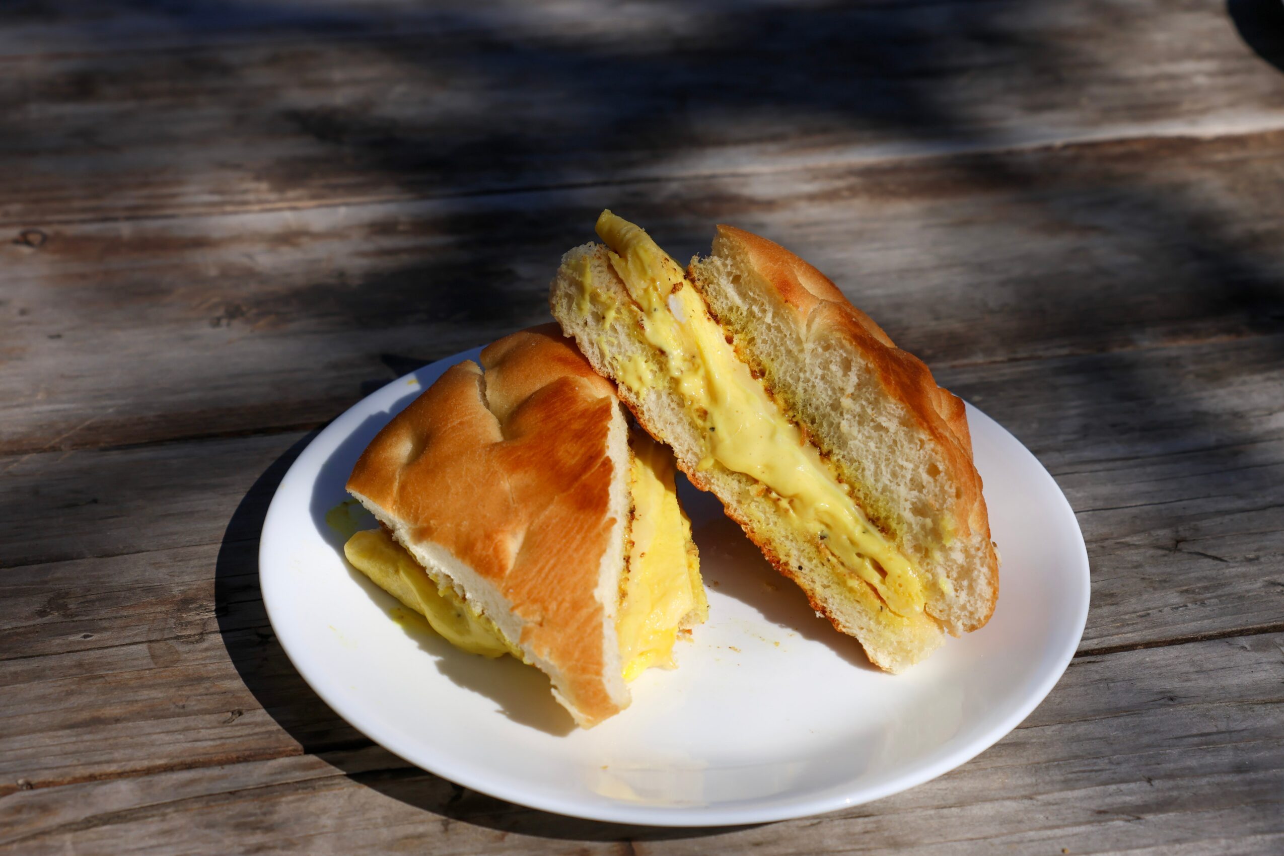A sandwich with egg filling on a white plate, placed on a wooden surface outdoors.