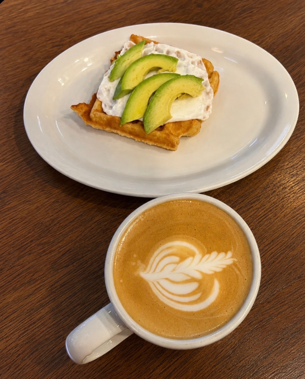 A plate with toast topped with cream and sliced avocado, and a cup of latte with latte art on a wooden table.