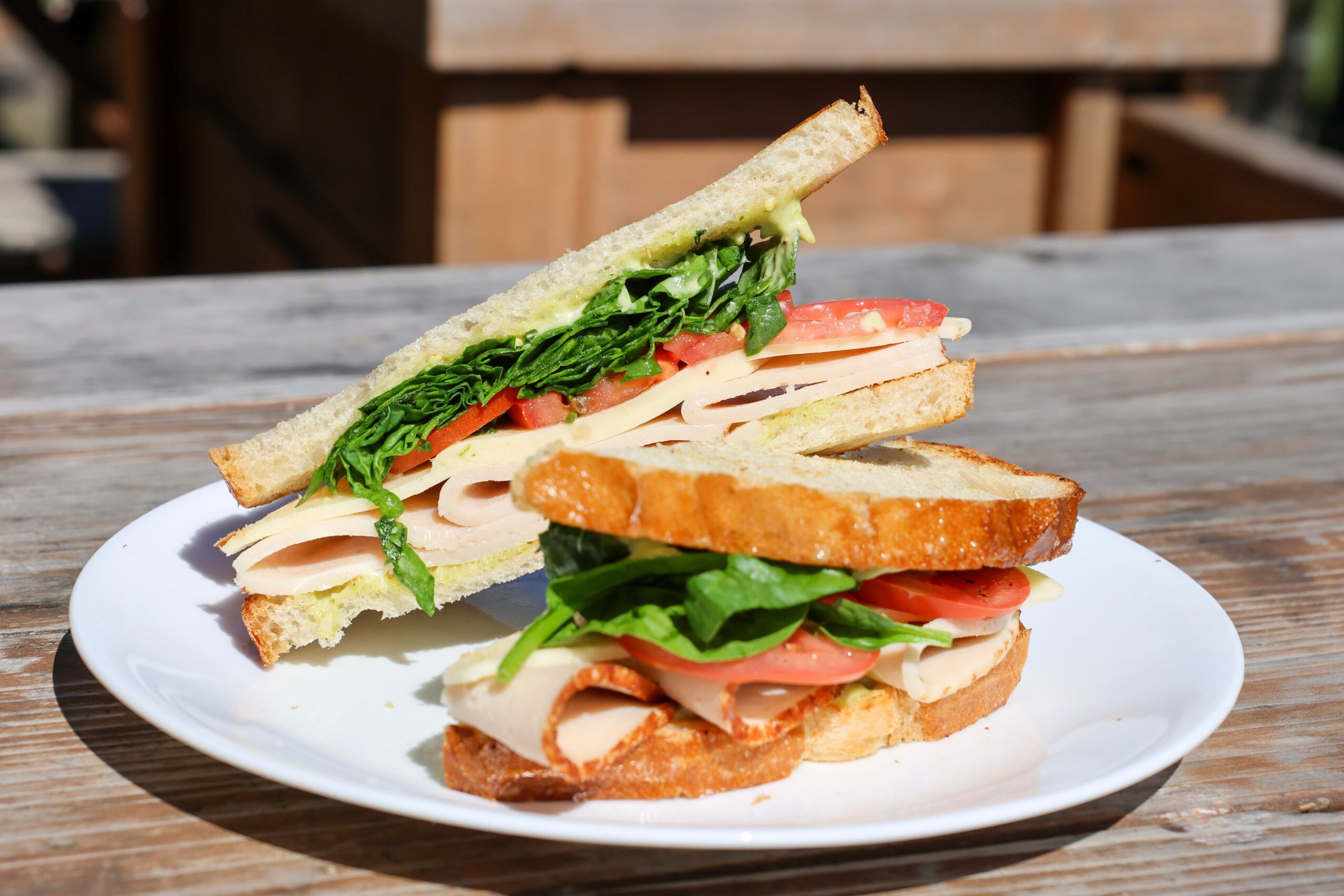 Sandwich with lettuce, tomato, turkey, and bacon on a white plate outdoors.