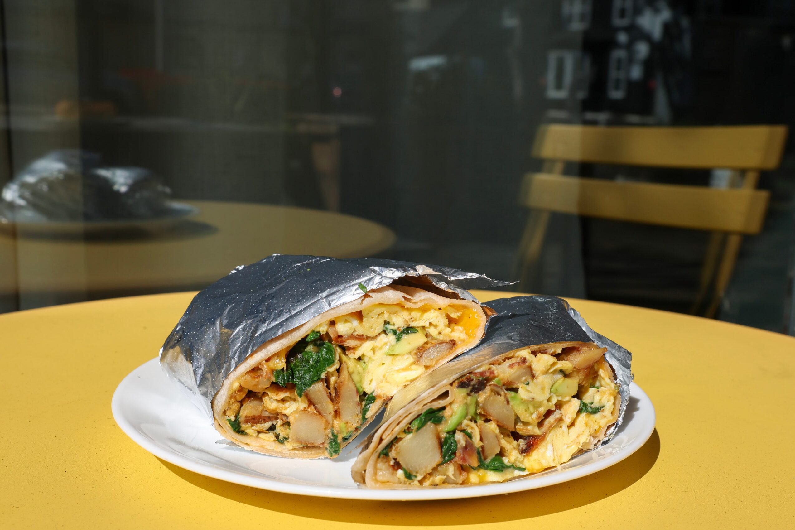 Two sushi burritos on a white plate, with seaweed wrapping and visible fillings of rice, vegetables, and possibly egg, on a yellow table.