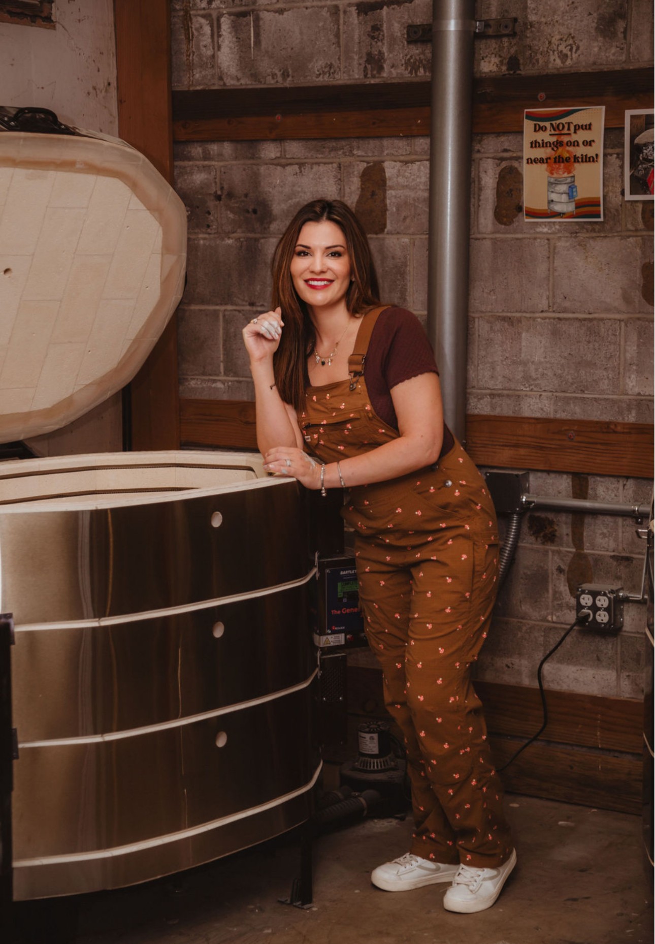 Young woman with long hair, smiling, standing indoors near a large metallic object, wearing a brown polka-dot jumpsuit and white sneakers.