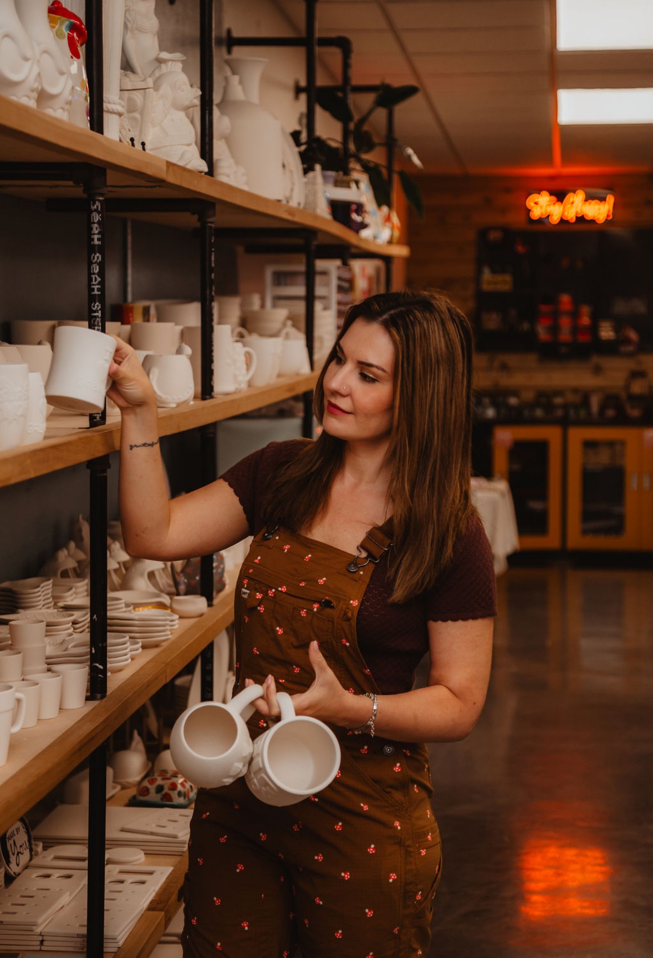 Woman shopping for white ceramic mugs on store shelves, holding two mugs in her hands, smiling, with shelves of ceramics behind her.