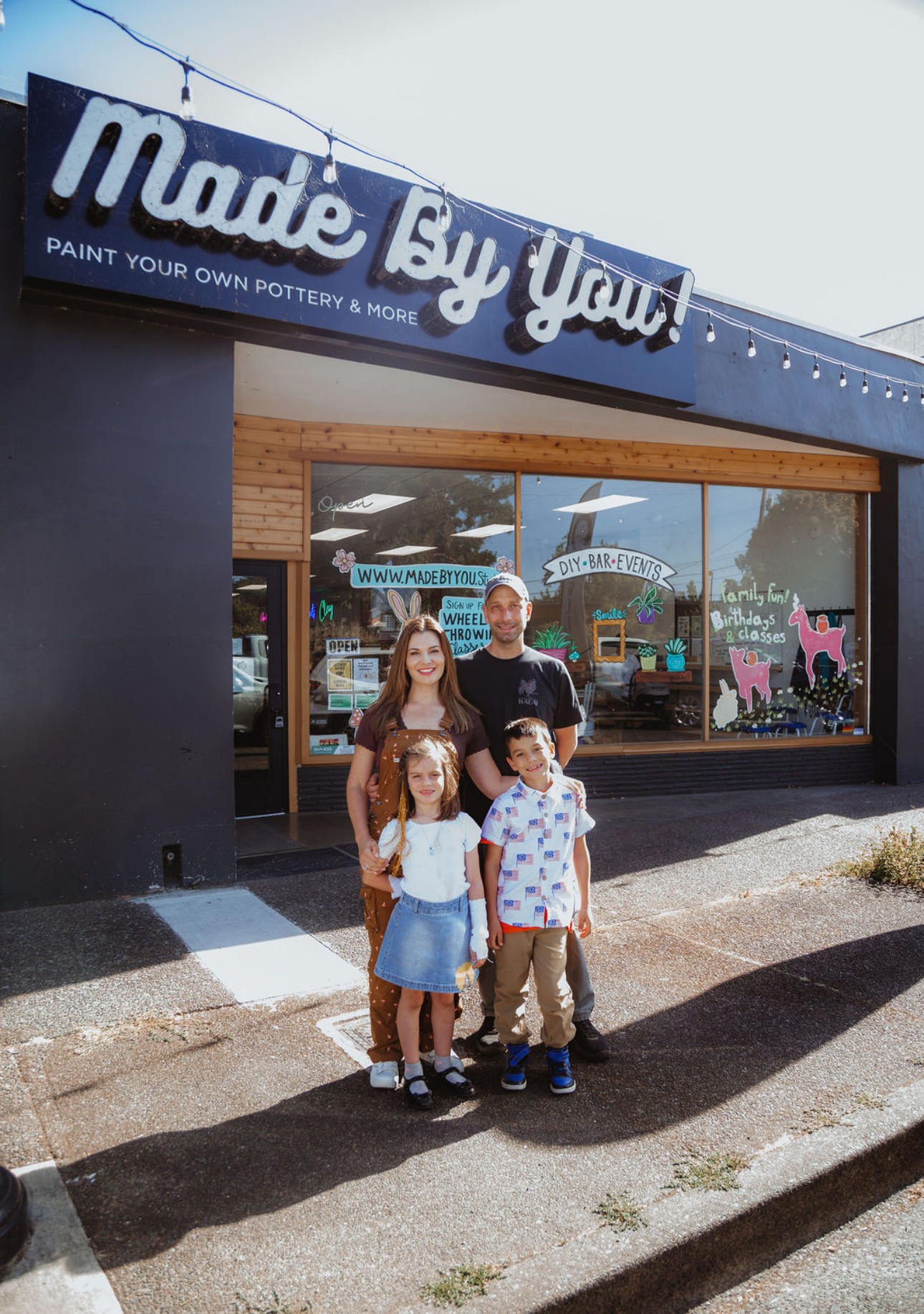 Four people, two adults and two children, stand outside a pottery painting shop with a sign that reads 'Made By You!'.
