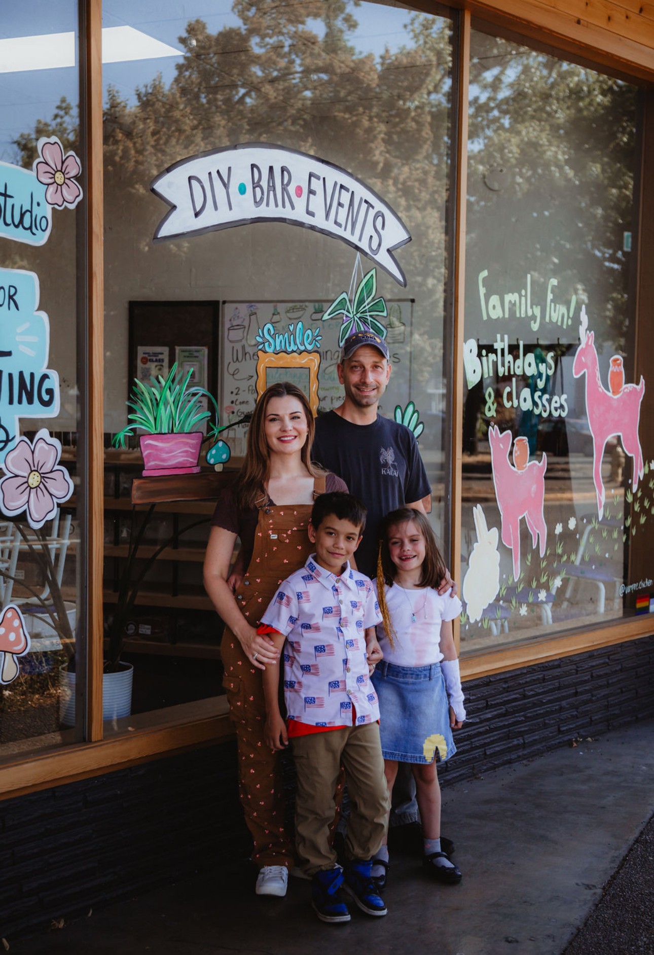 Family of four standing outside a decorated shop window with signs for DIY, bar, and events, smiling at the camera.