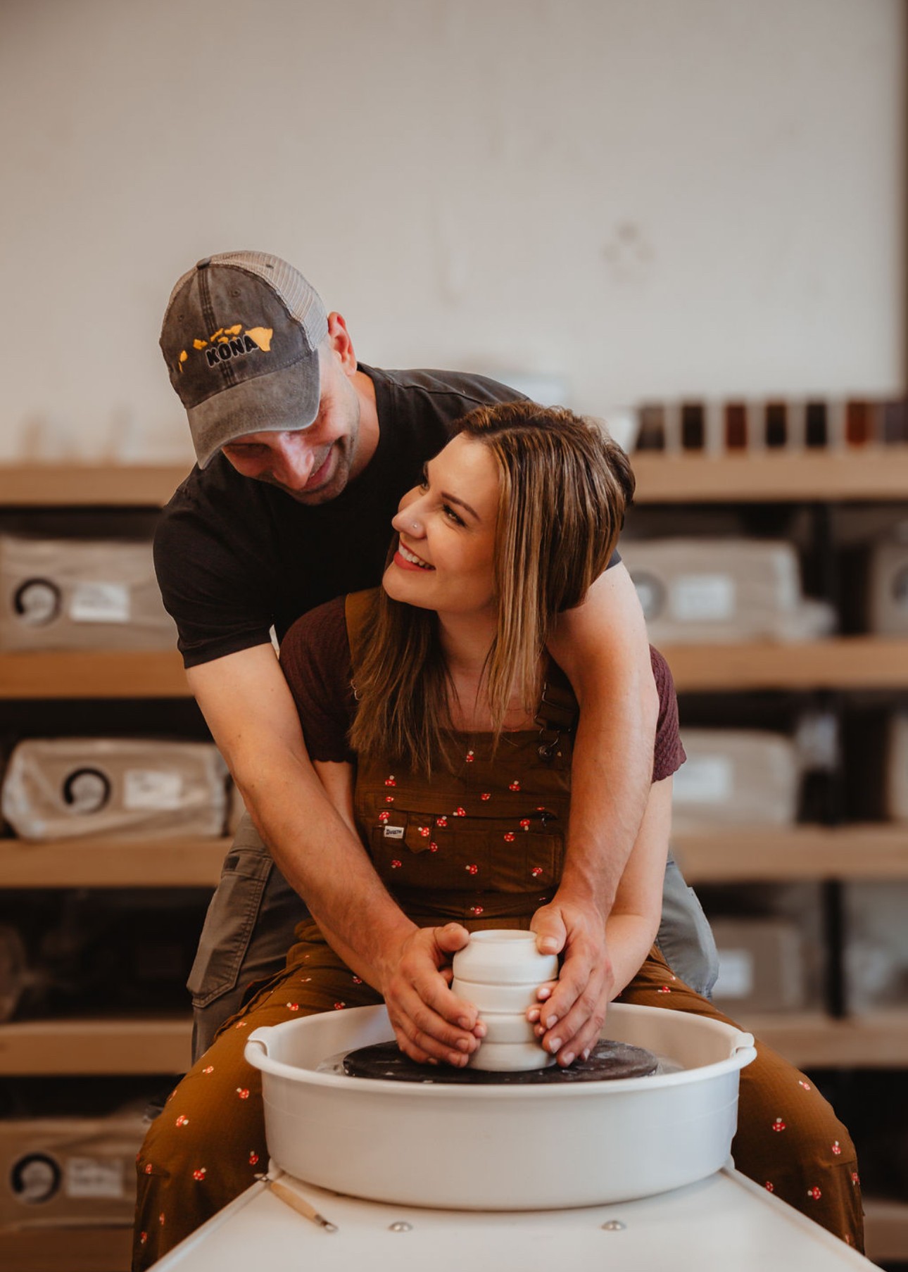 Two people, a man and a woman, sit at a pottery wheel, shaping clay together, smiling at each other.