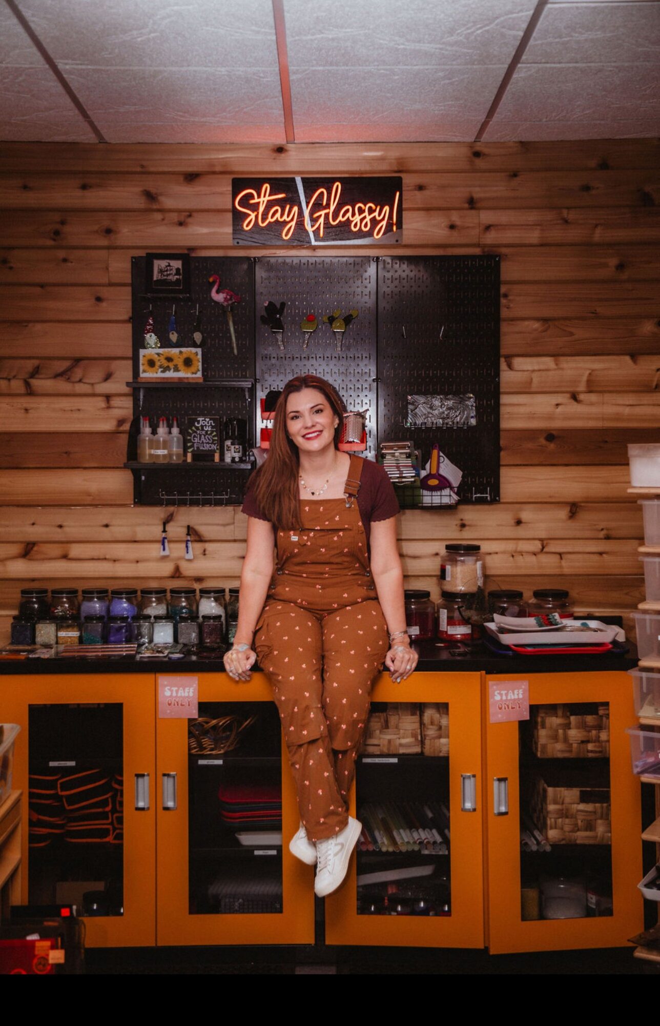 Woman standing in front of a black wall with shelves, smiling, wearing brown polka dot outfit and white shoes, wooden wall background.