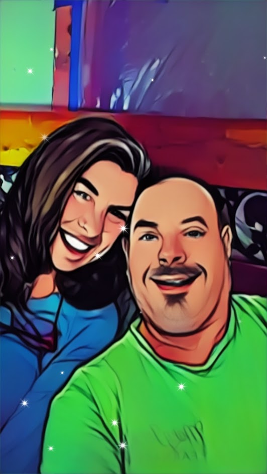Two smiling people, a woman with dark hair and a man with a shaved head, pose together indoors.