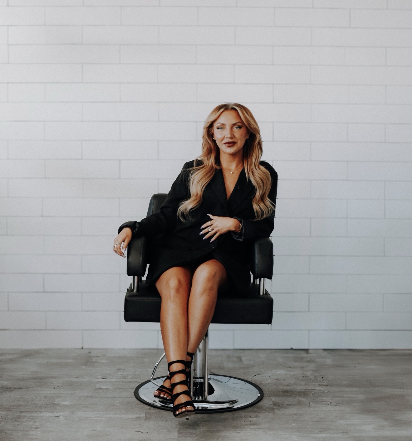Woman with long blonde hair sitting on a black chair against a white brick wall, wearing a black dress and strappy heels.