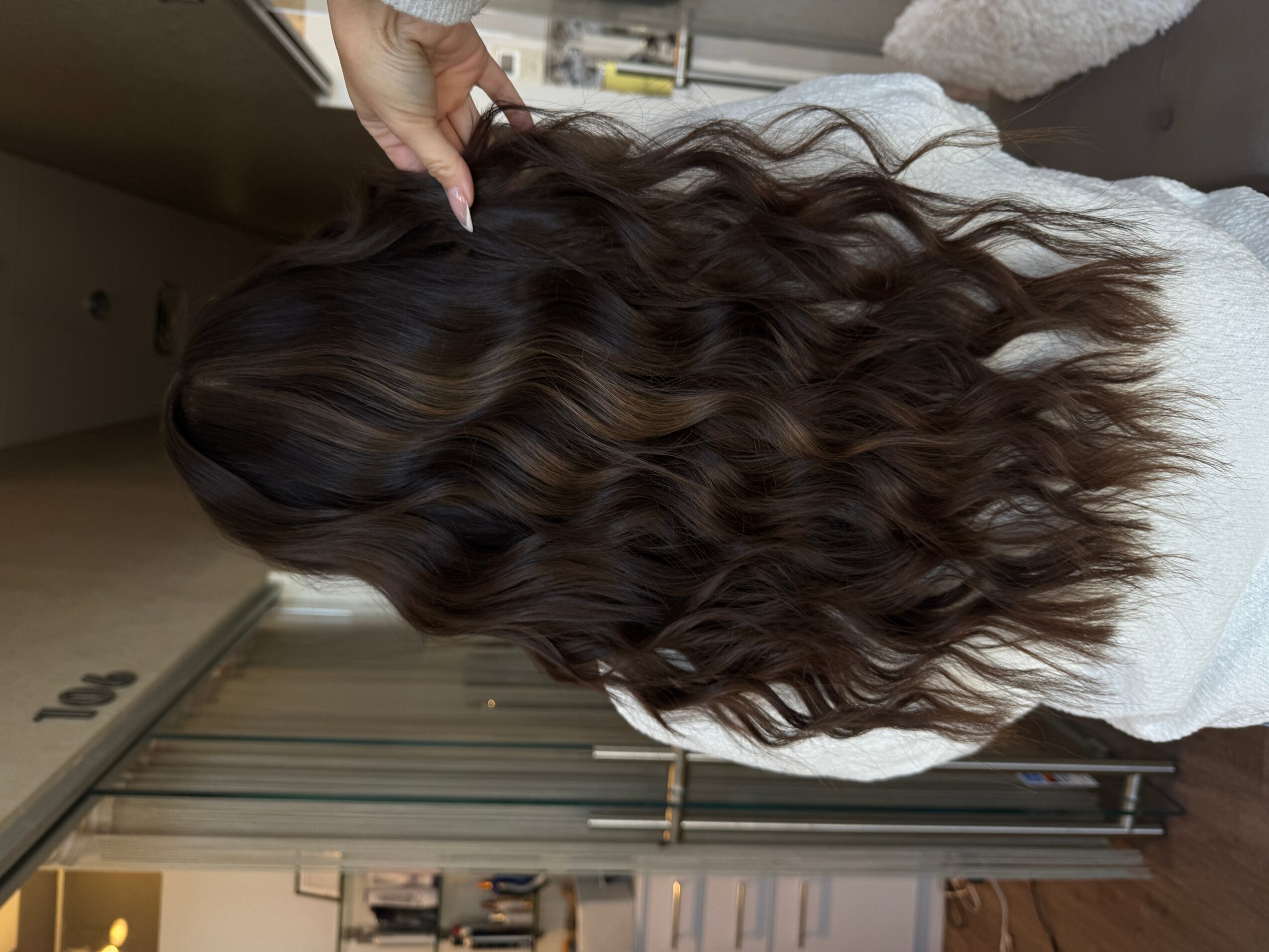 Person with long, wavy brown hair, viewed from behind, holding a section of hair. Indoor setting with cabinets and curtains.