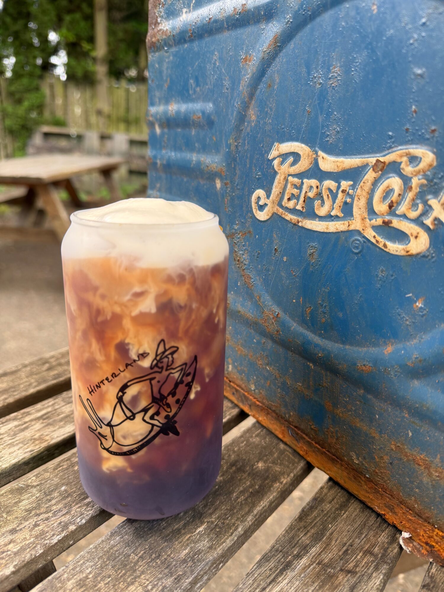 A colorful beverage in a glass with a logo, placed on a wooden surface next to a blue Pepsi cooler.