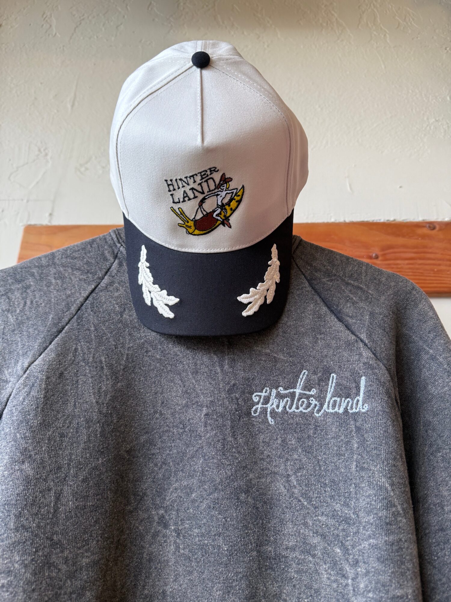 Cap with a white top, black brim, and embroidered design, placed on a gray sweatshirt with white text, on a wooden surface.