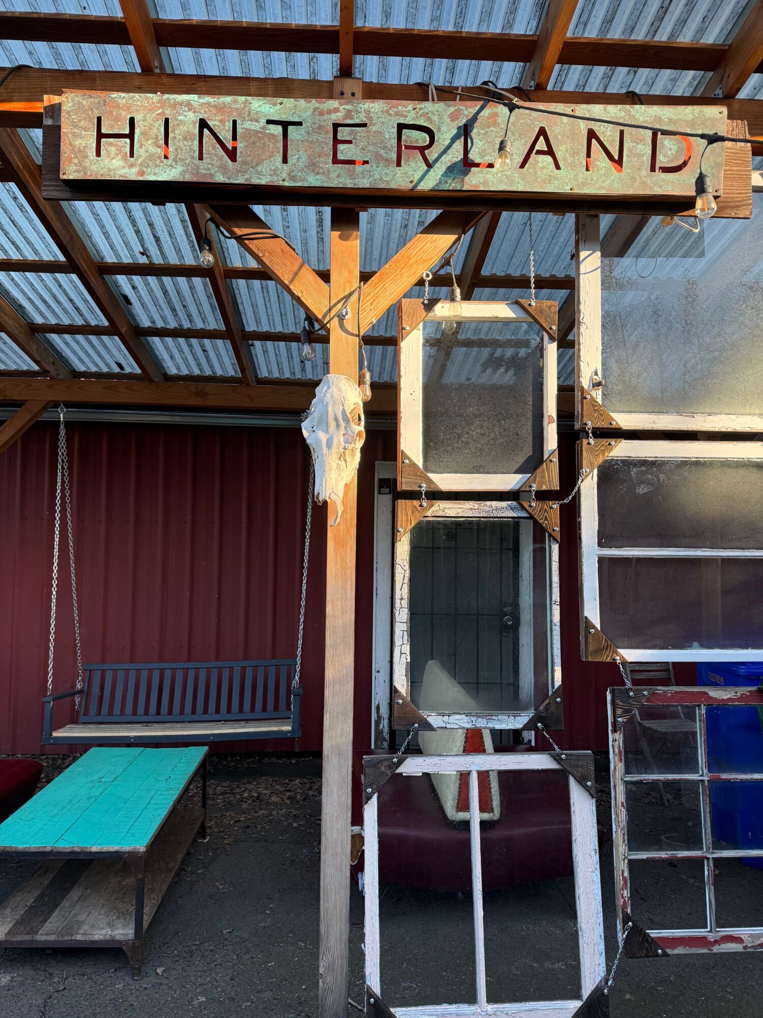 Wooden sign with the word 'HINTERLAND' hanging under a metal roof, with a bench and glass display cases nearby.