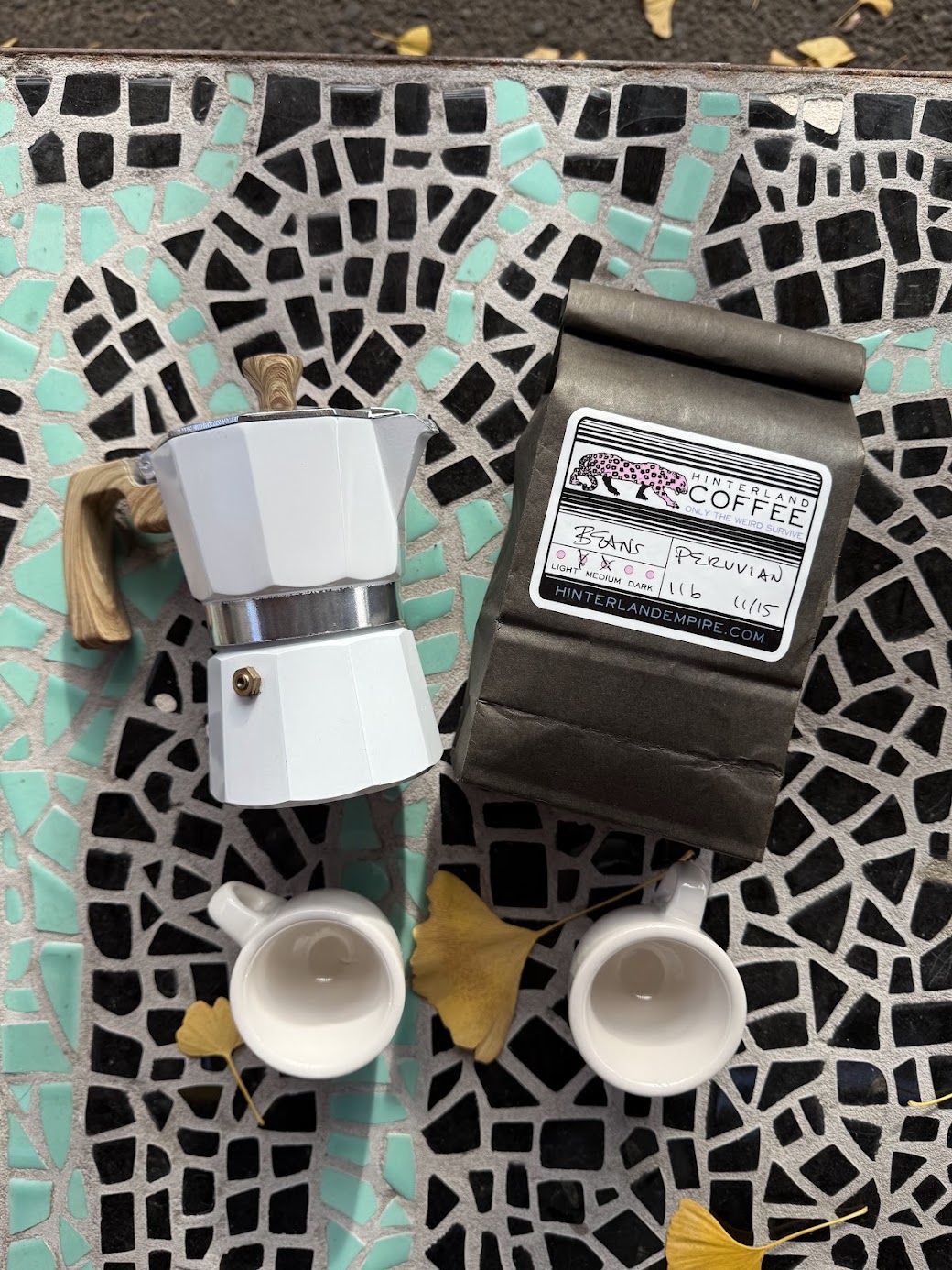 Two coffee cups with a coffee machine and a black wallet on a mosaic surface with yellow leaves, top view.