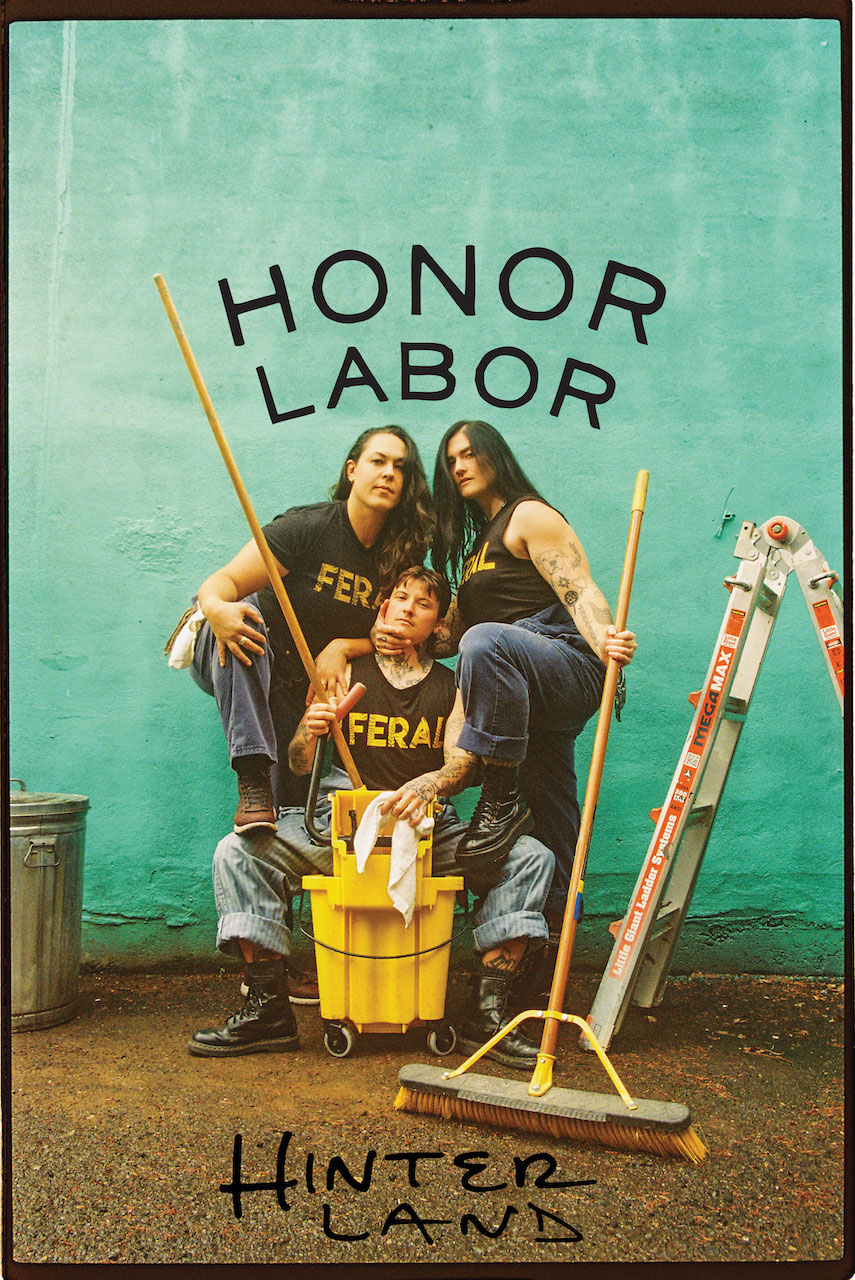 Three women in yellow shirts with 'FERA' text pose with cleaning tools against a teal wall with 'HONOR LABOR' and 'HINTER LAND' written on it.