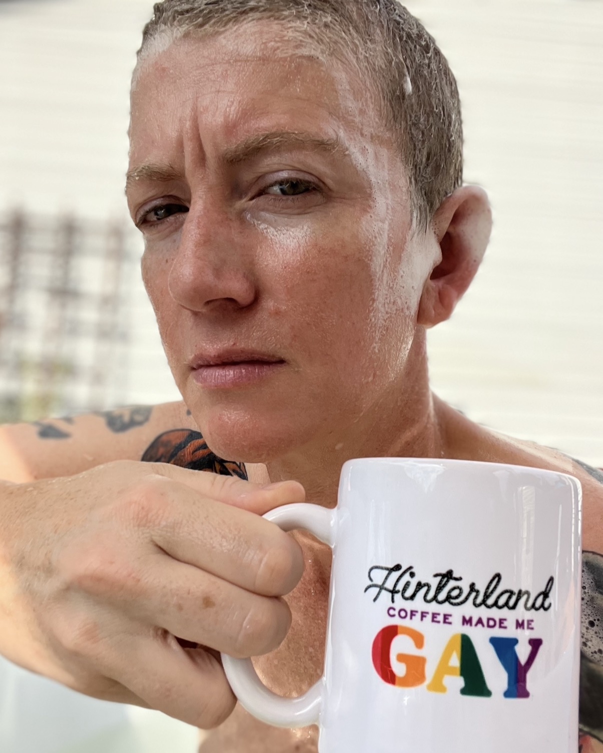 Person with short hair holding a white mug with colorful text, outdoors background, serious expression.