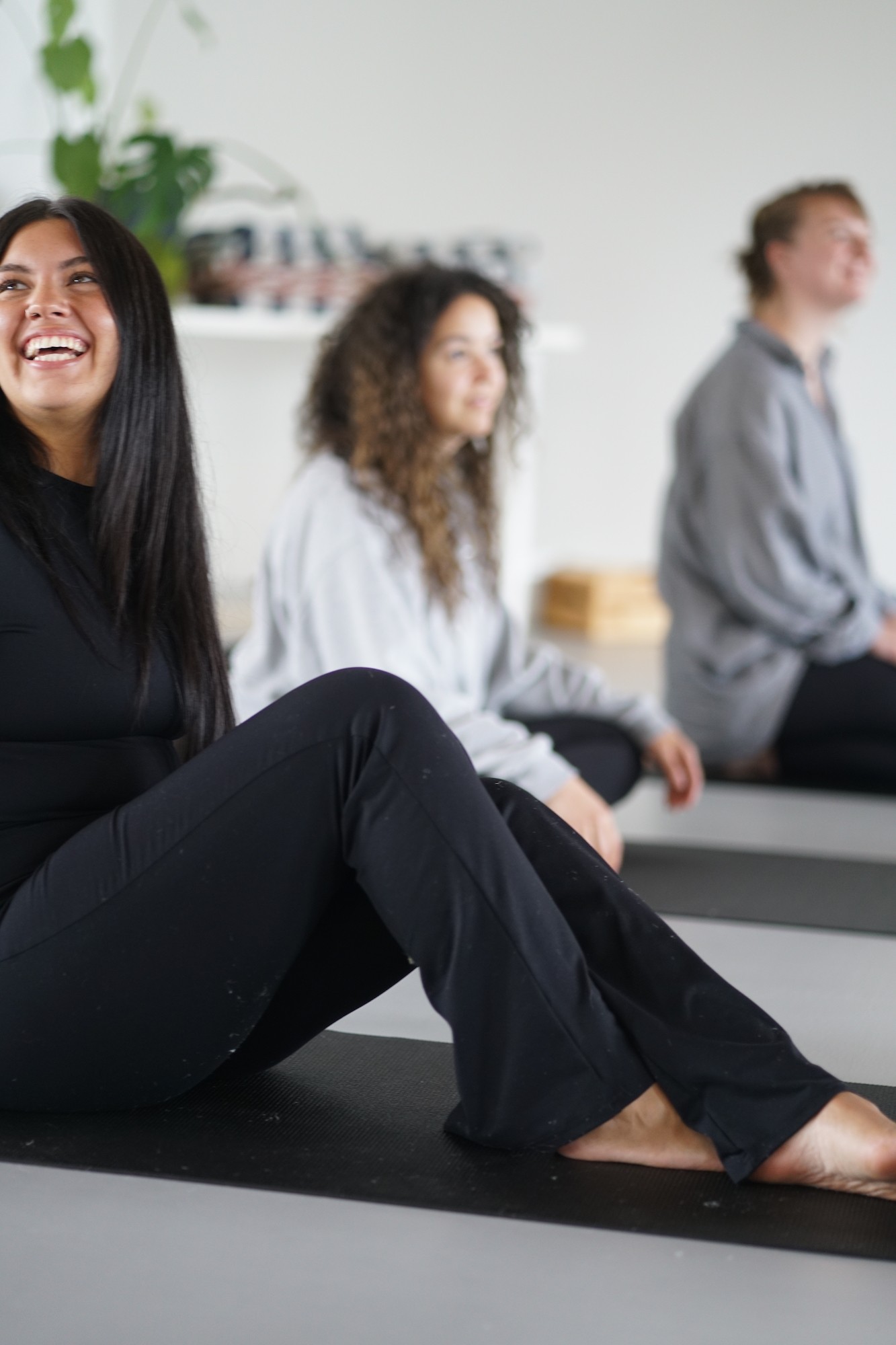 Three people sitting on a yoga mat, smiling and relaxed in a bright room with plants and shelves.