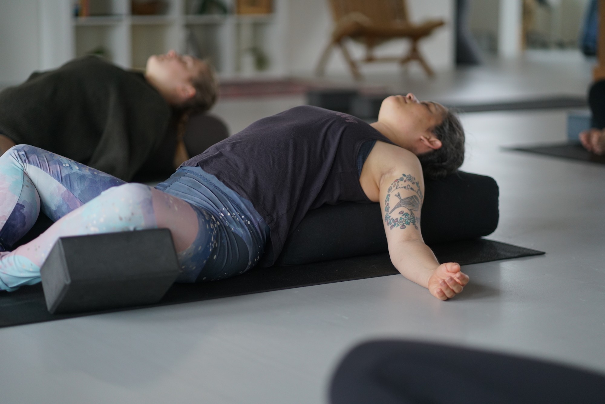 Two people lying on the floor, one on a yoga mat with foam blocks, in a room with chairs and shelves.