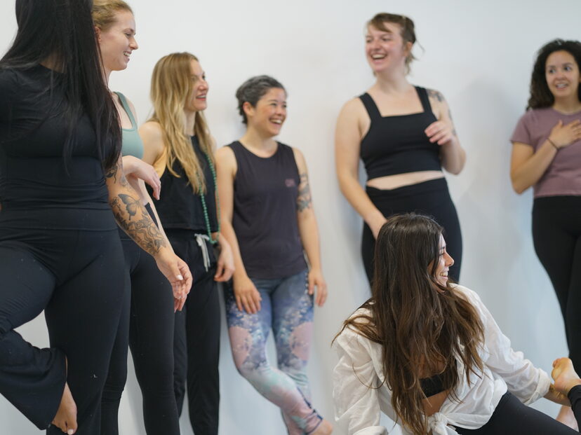 Group of six women standing and laughing together in a bright room, some with tattoos and casual clothing.