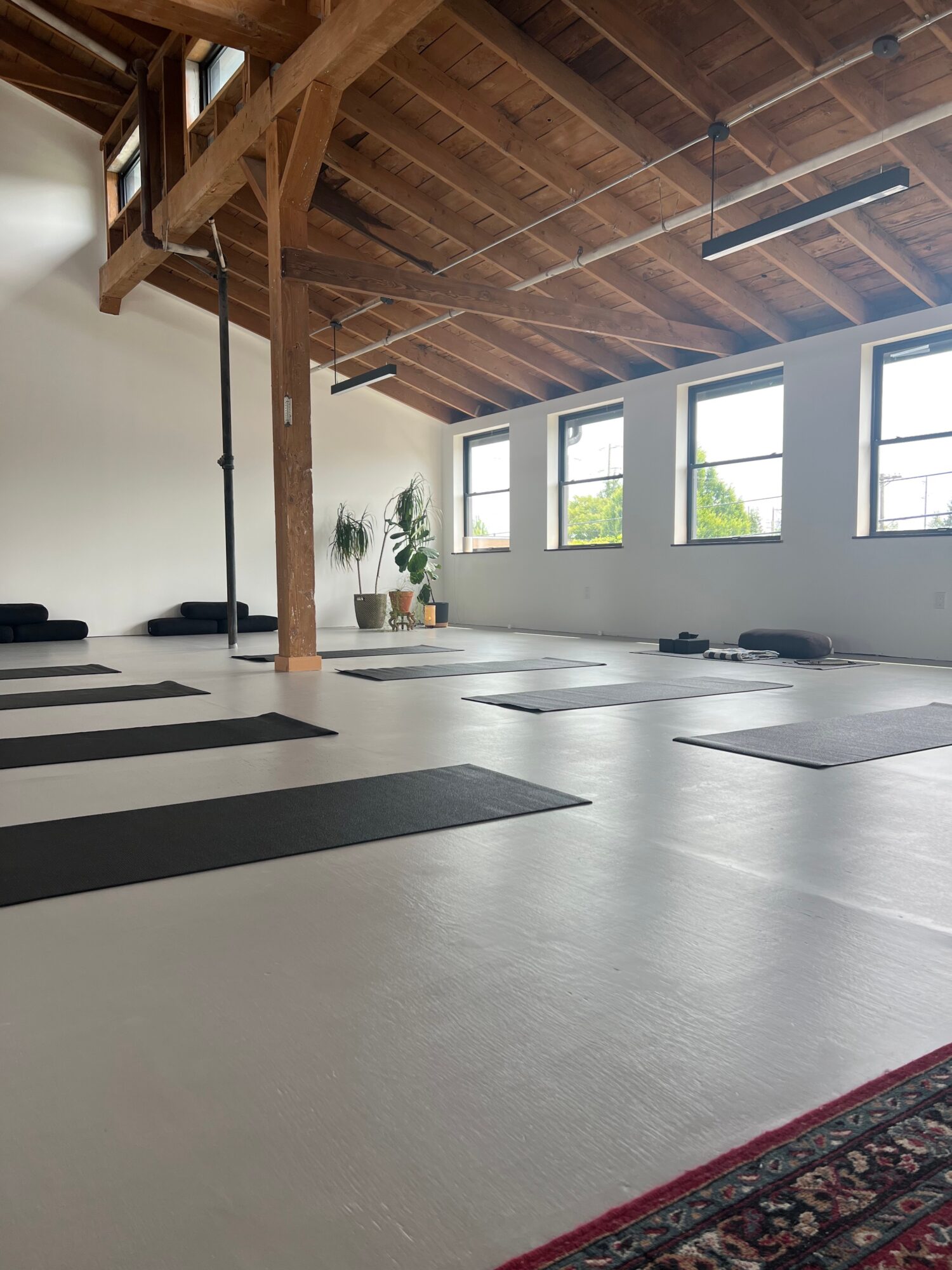 Yoga mats arranged on a spacious, well-lit studio with large windows, wooden ceiling, and minimal decor.