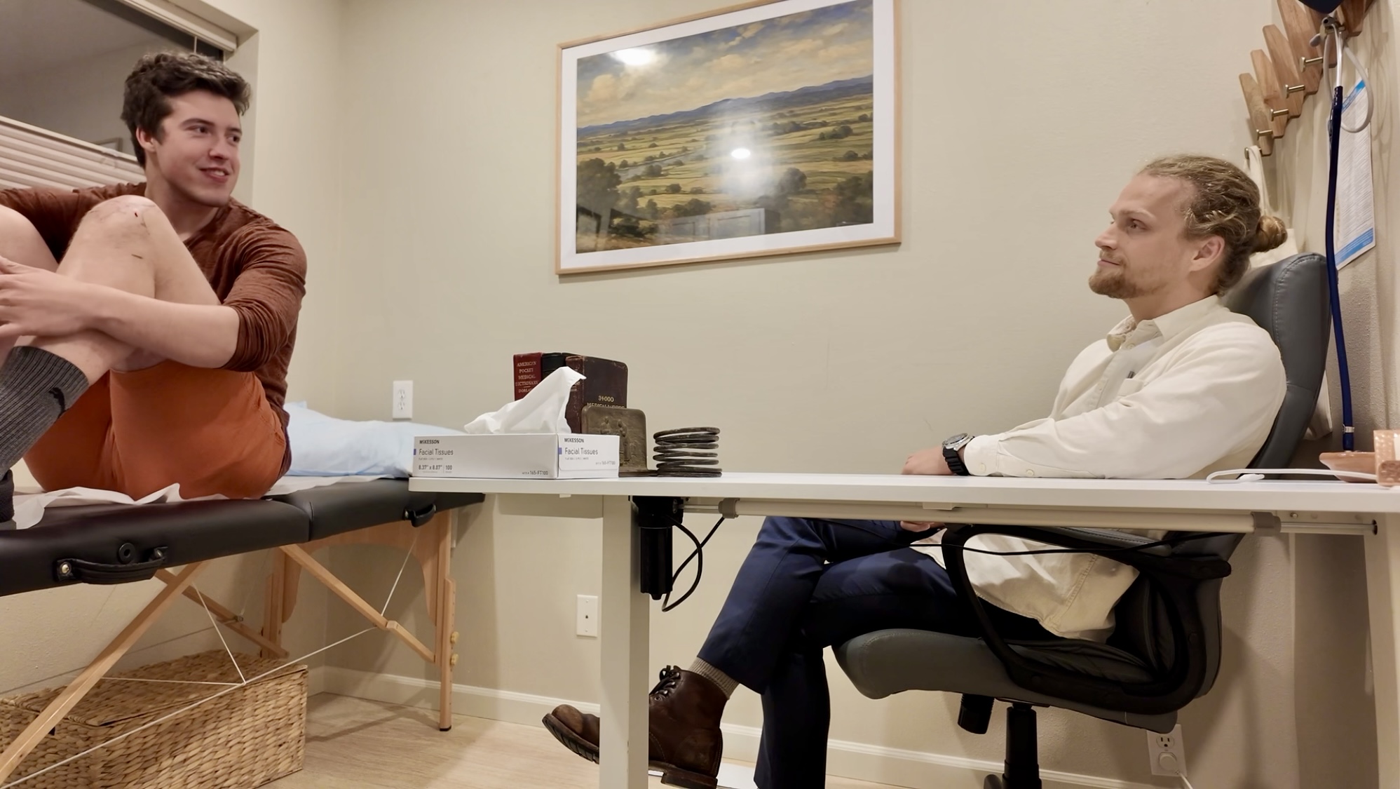 A person lying on a table with legs raised, talking to a seated person in an office or therapy room, with a painting on the wall.