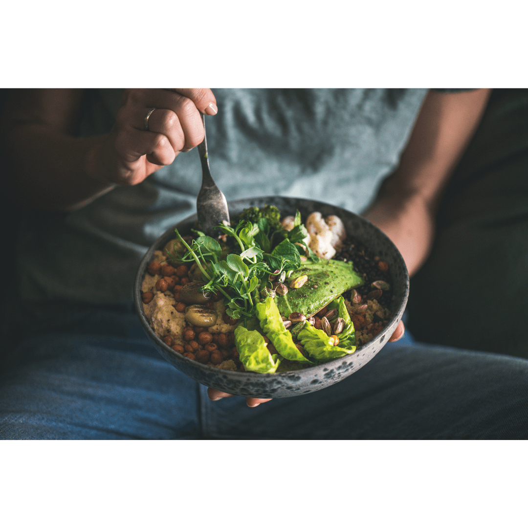 Person holding a bowl of salad with greens and vegetables, using a fork, seated with a gray shirt and blue jeans.