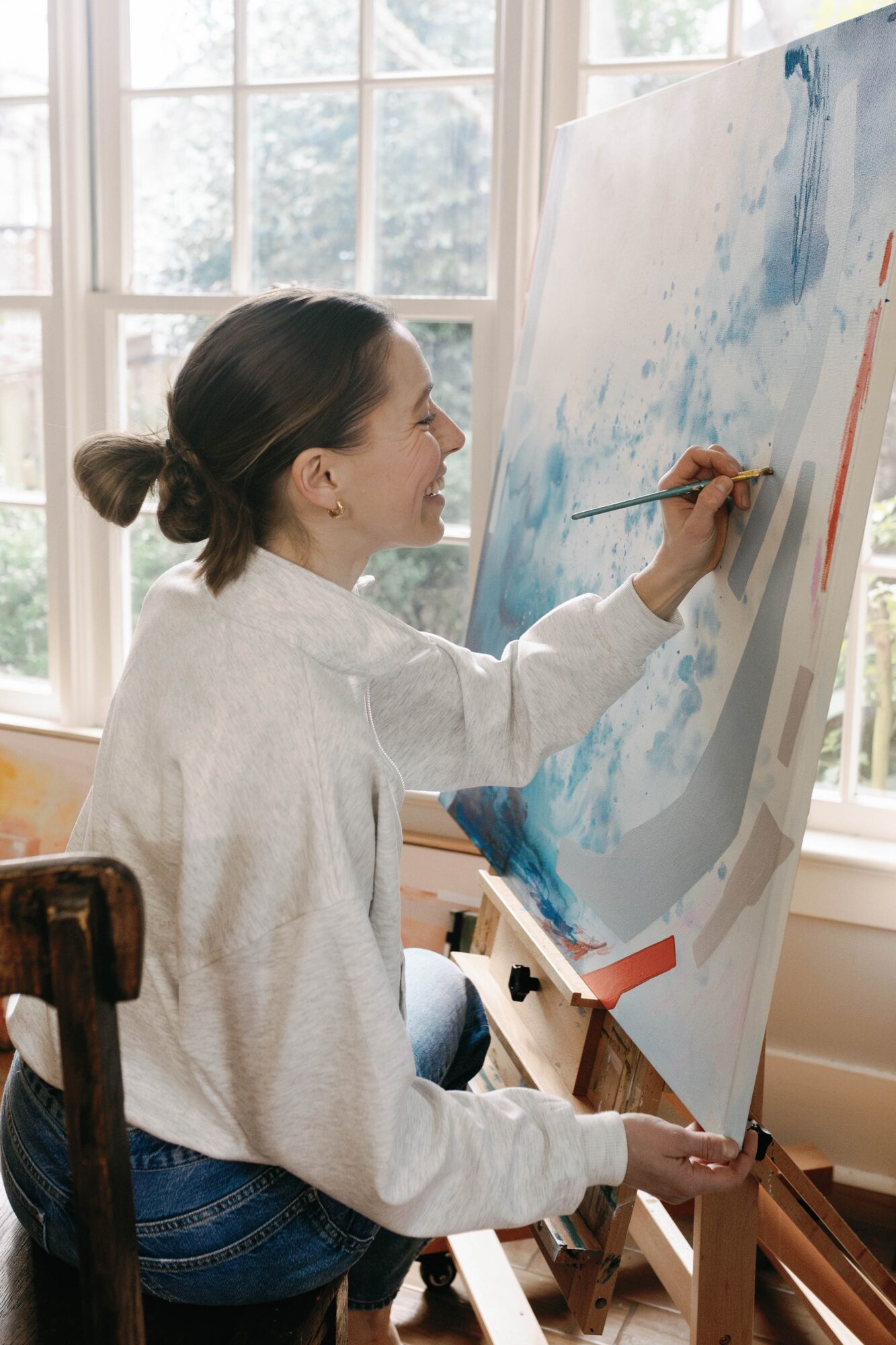 Person painting on large canvas in bright room with large windows, wearing white sweater, holding paintbrush and palette.