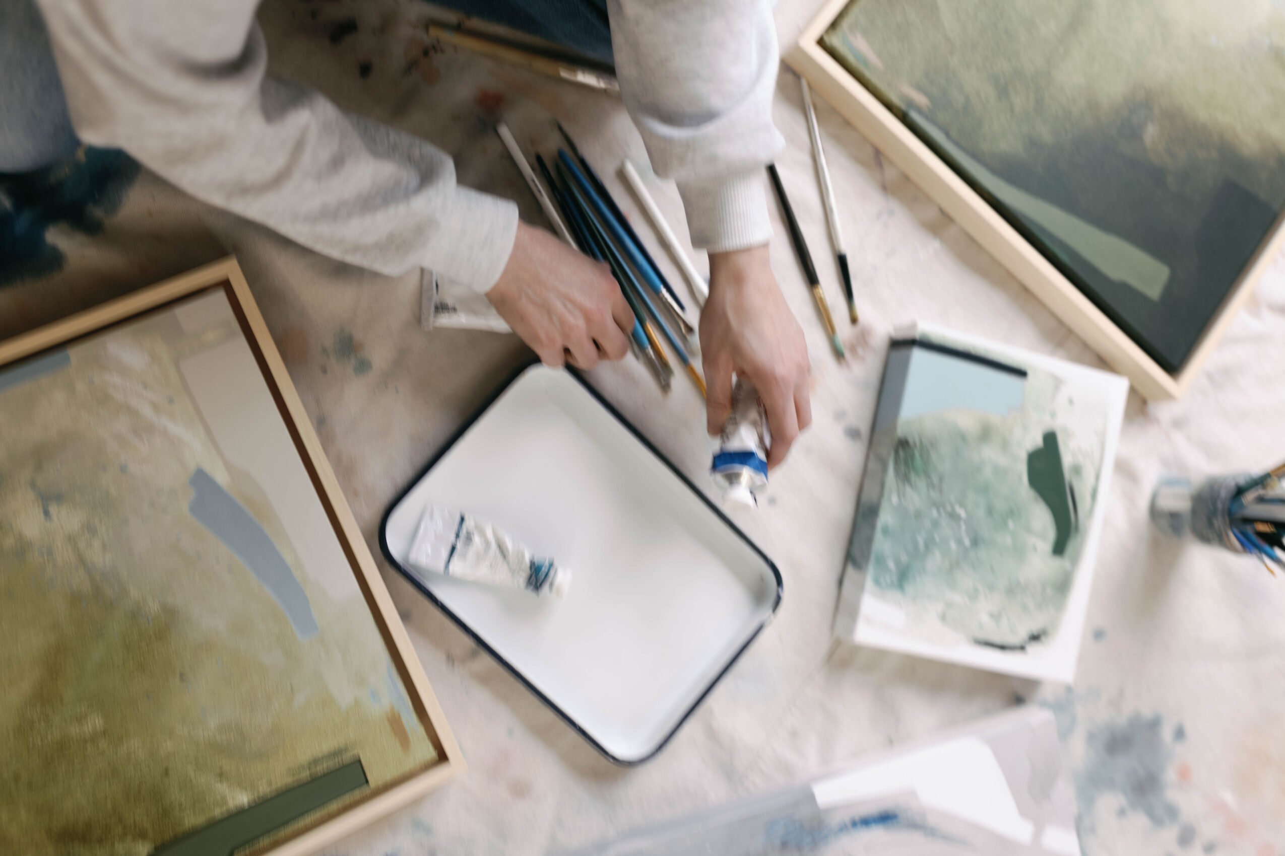 Person arranging art supplies on table with framed paintings and brushes, viewed from above.