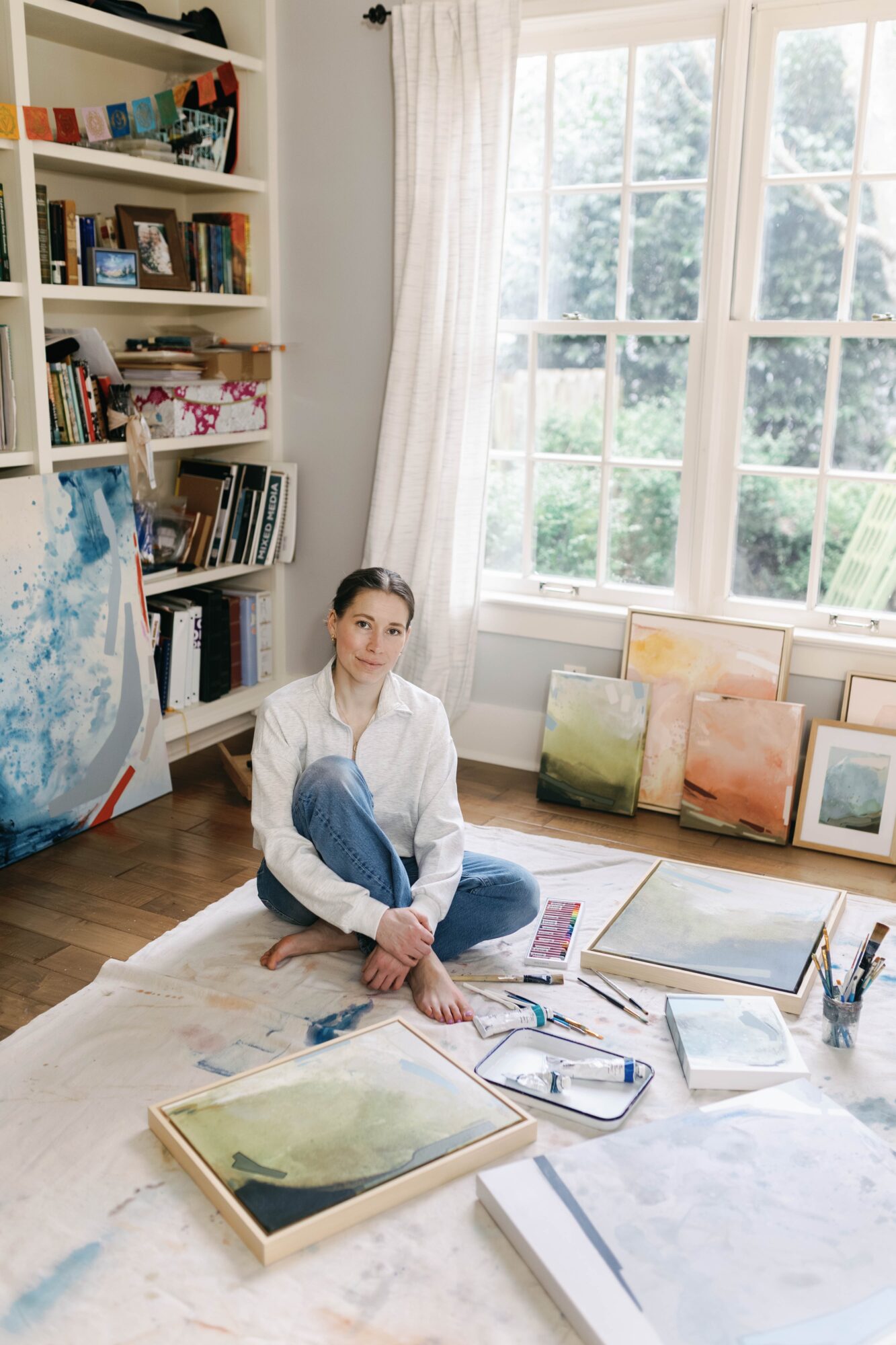 Woman sitting on the floor in an art studio with paintings, books, and large windows behind her.