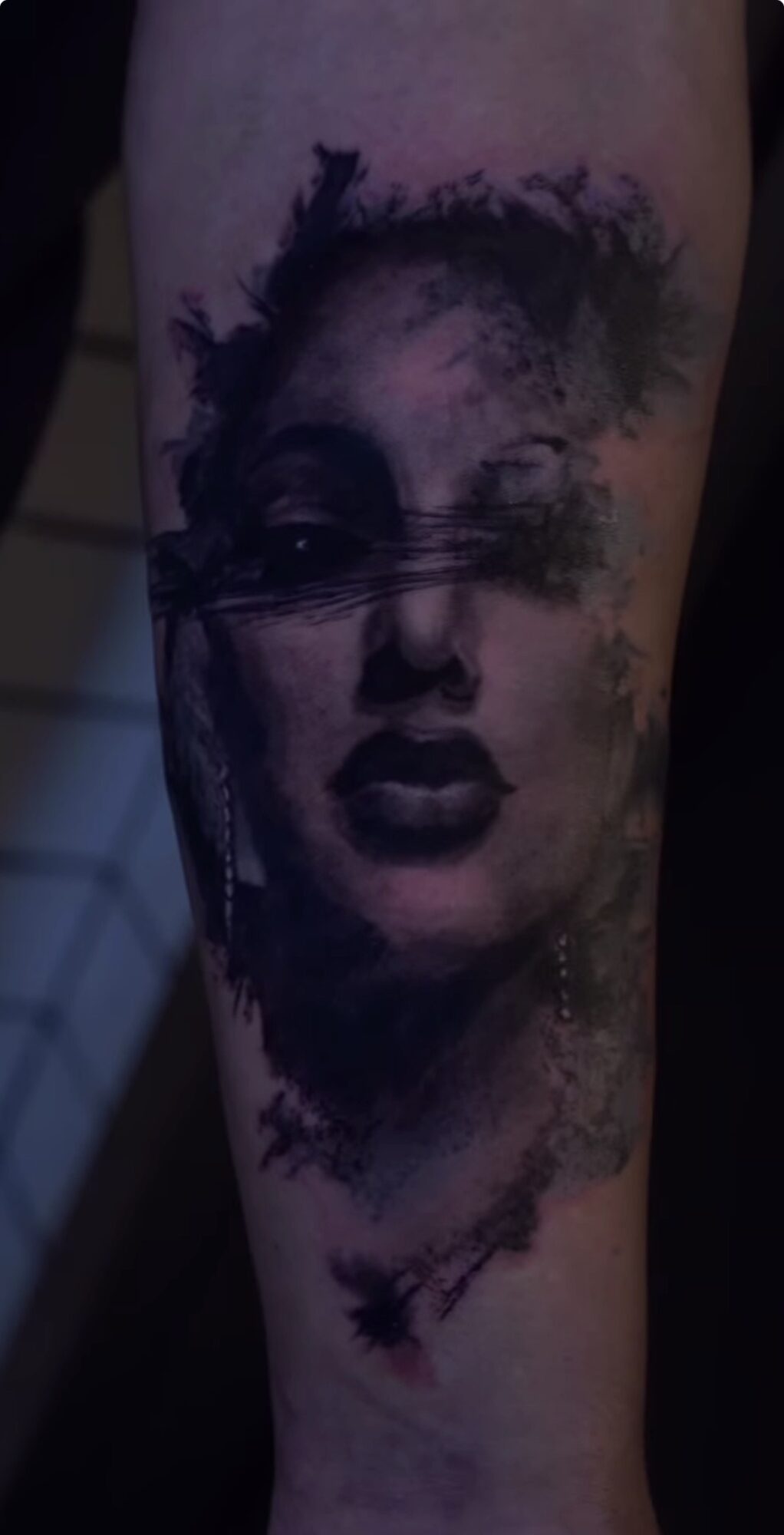 Tattoo of a woman's face with dark shading and abstract elements on skin.