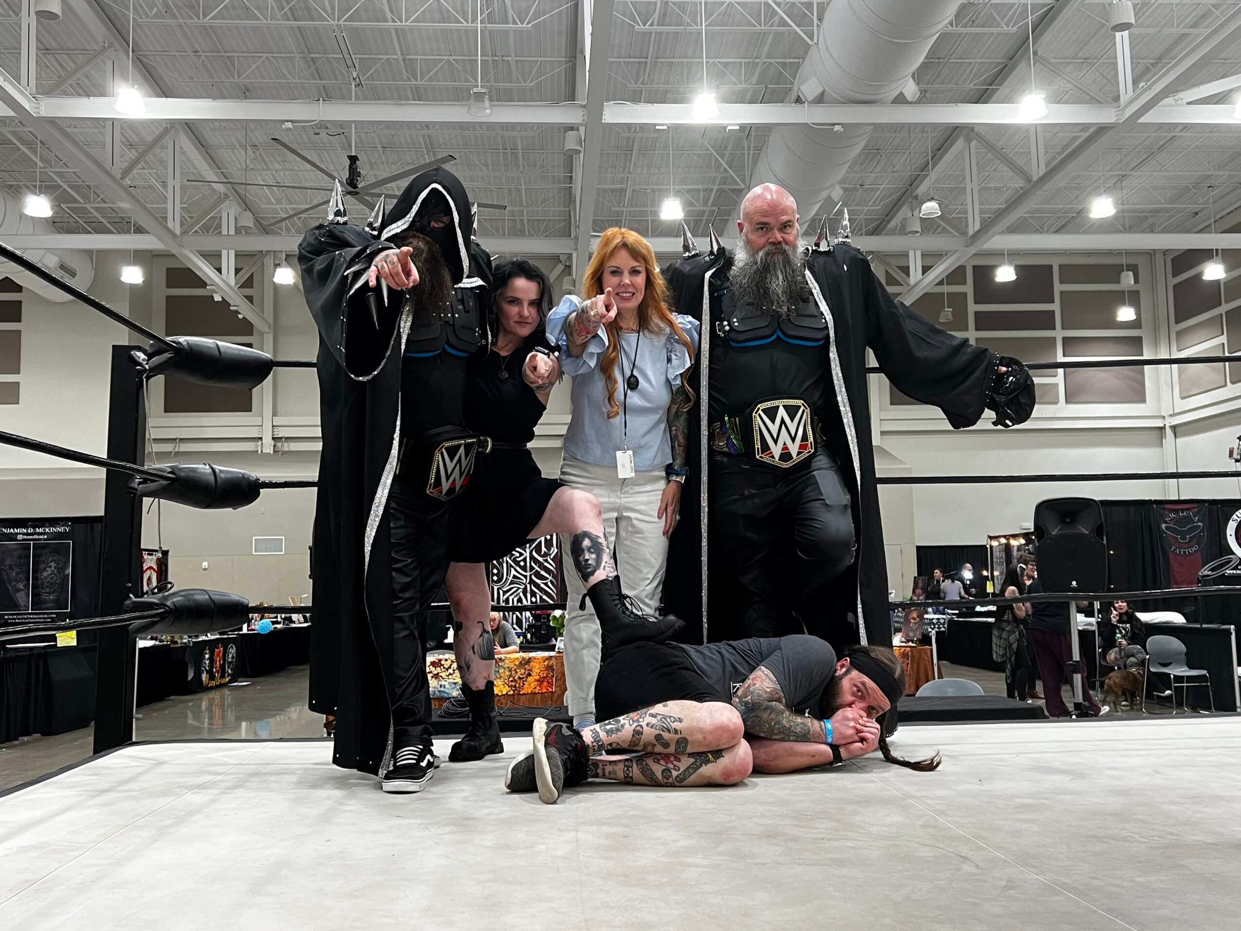 Four people in wrestling costumes and casual clothing pose in a wrestling ring, with one person lying on the floor. The background shows a large indoor space with equipment and people.