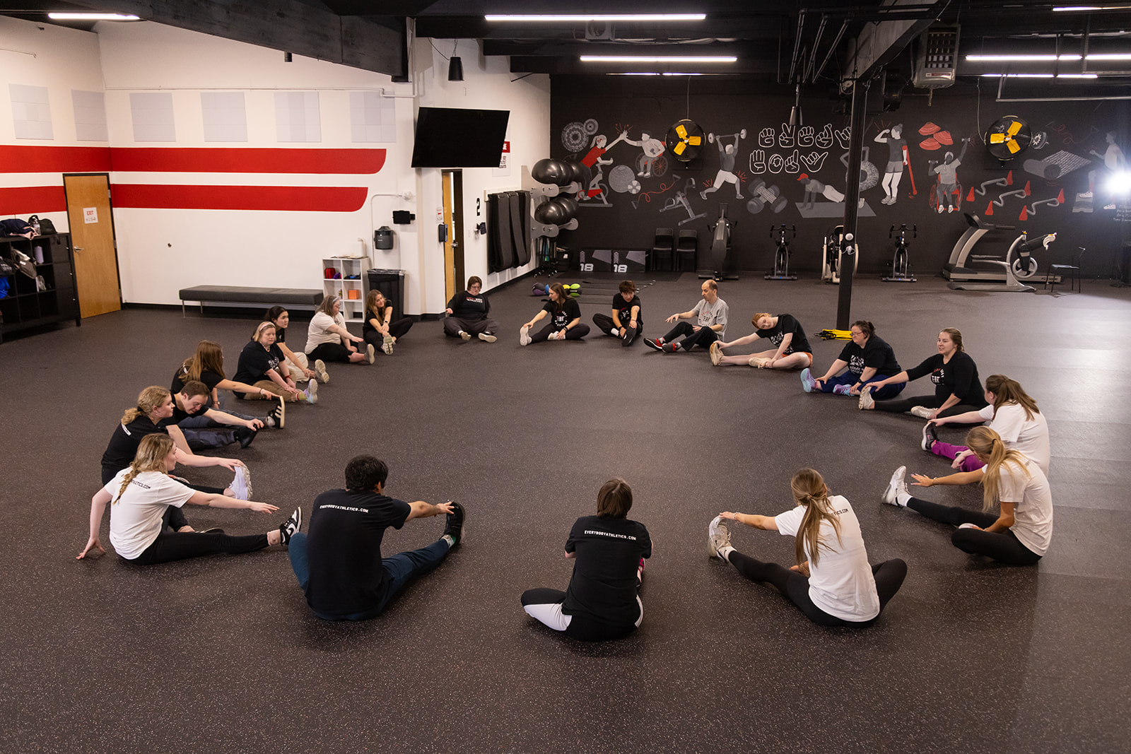 Group of people sitting in a circle on a gym floor, participating in a fitness class or activity.