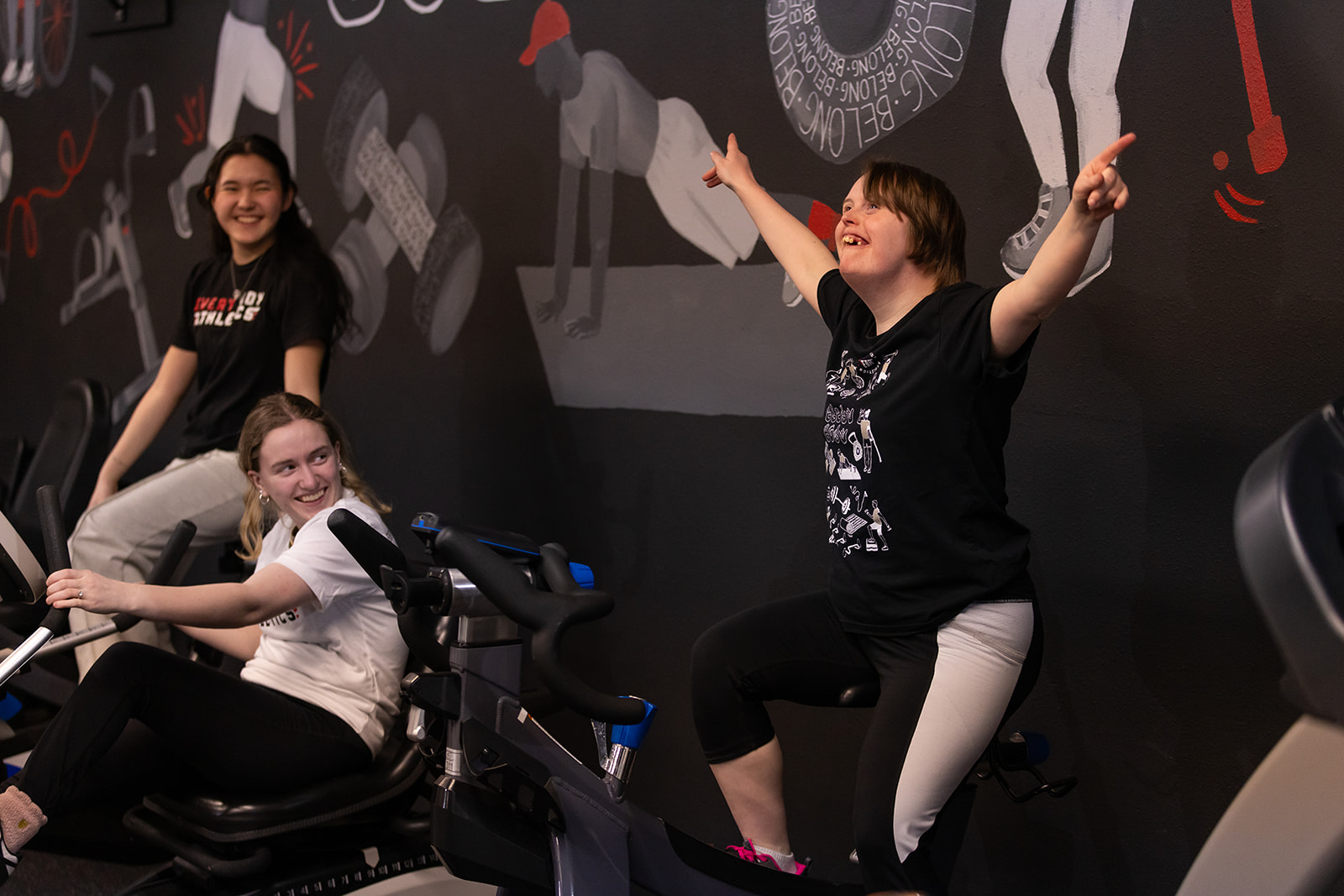 Three people in a gym, one on a stationary bike, two standing and smiling, with a mural on the wall behind them.