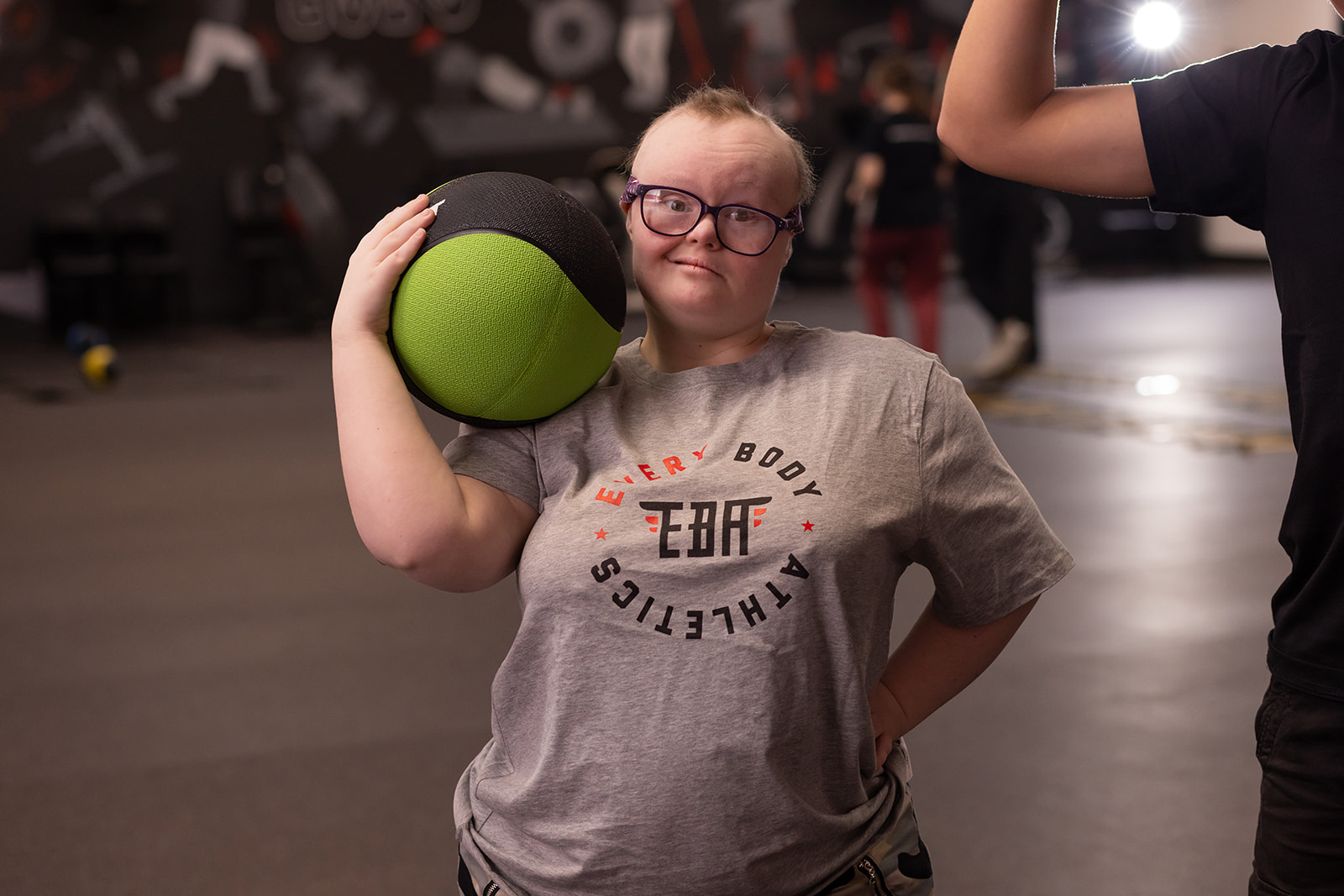 Person holding a green and black medicine ball on shoulder, wearing glasses and a gray T-shirt, in a gym.