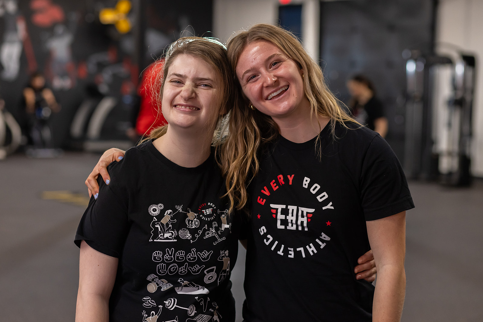 Two smiling women with arms around each other in a gym, wearing black t-shirts, background with gym equipment.