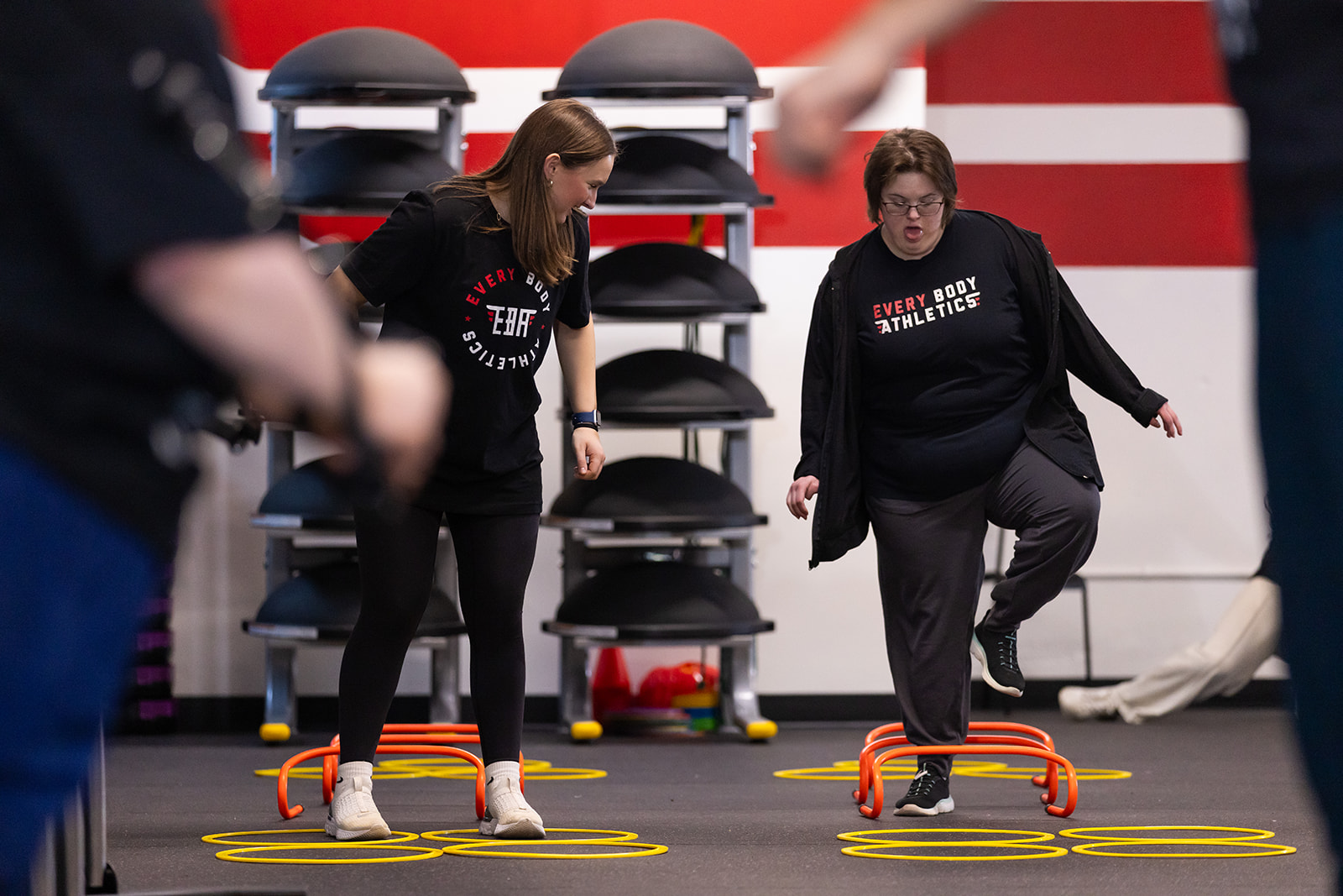 Two women participate in an indoor fitness activity, stepping over small hurdles on a black floor with yellow circles, in a gym setting.