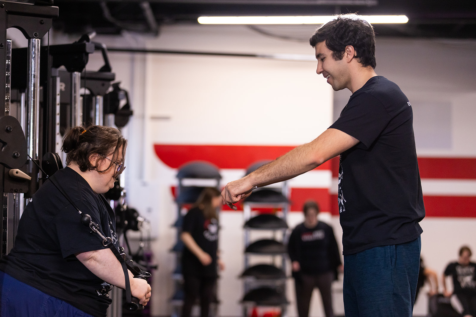 A young man and a young woman interact in a gym, with other people and gym equipment in the background.