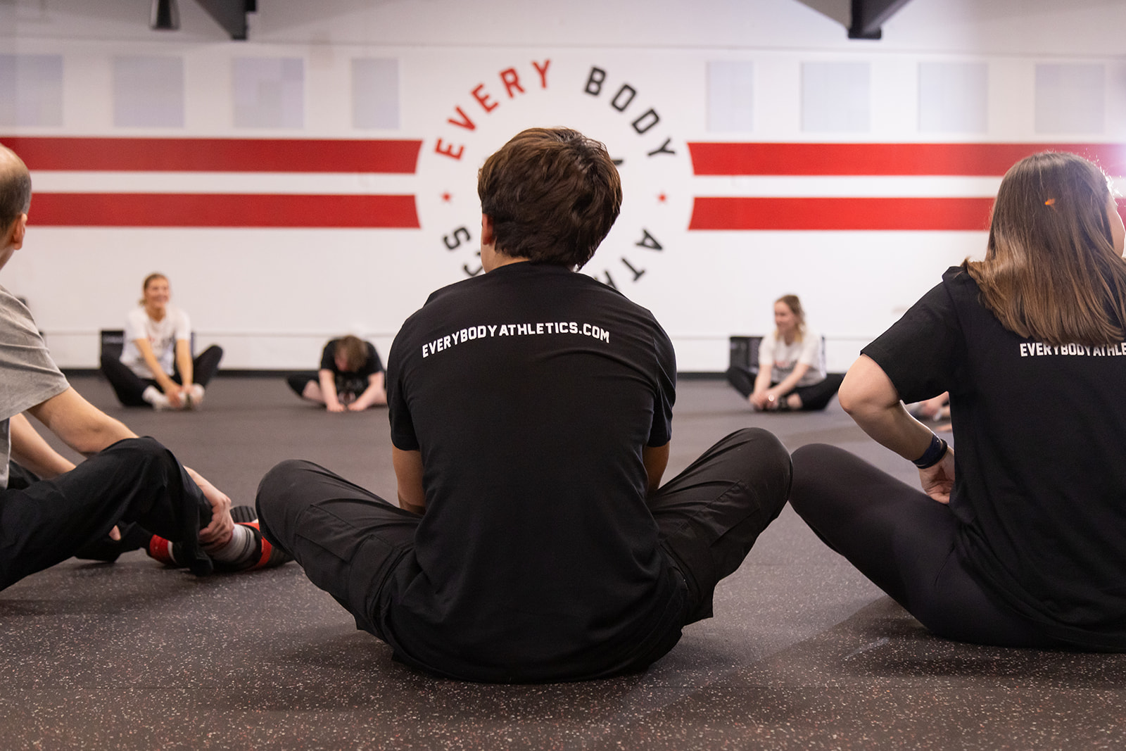 Group of people sitting on the floor in a gym, facing a wall with a logo and text, some stretching or resting.