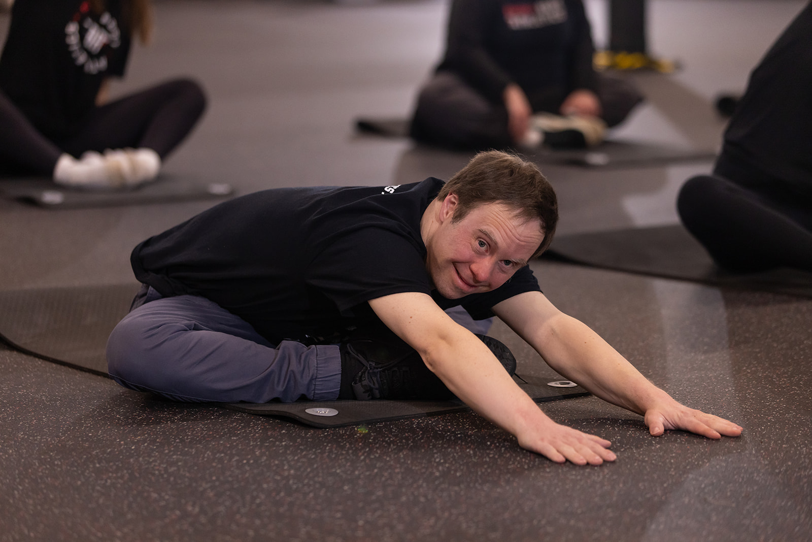 Person stretching on a mat in a group exercise class, lying forward with arms extended, smiling at the camera.