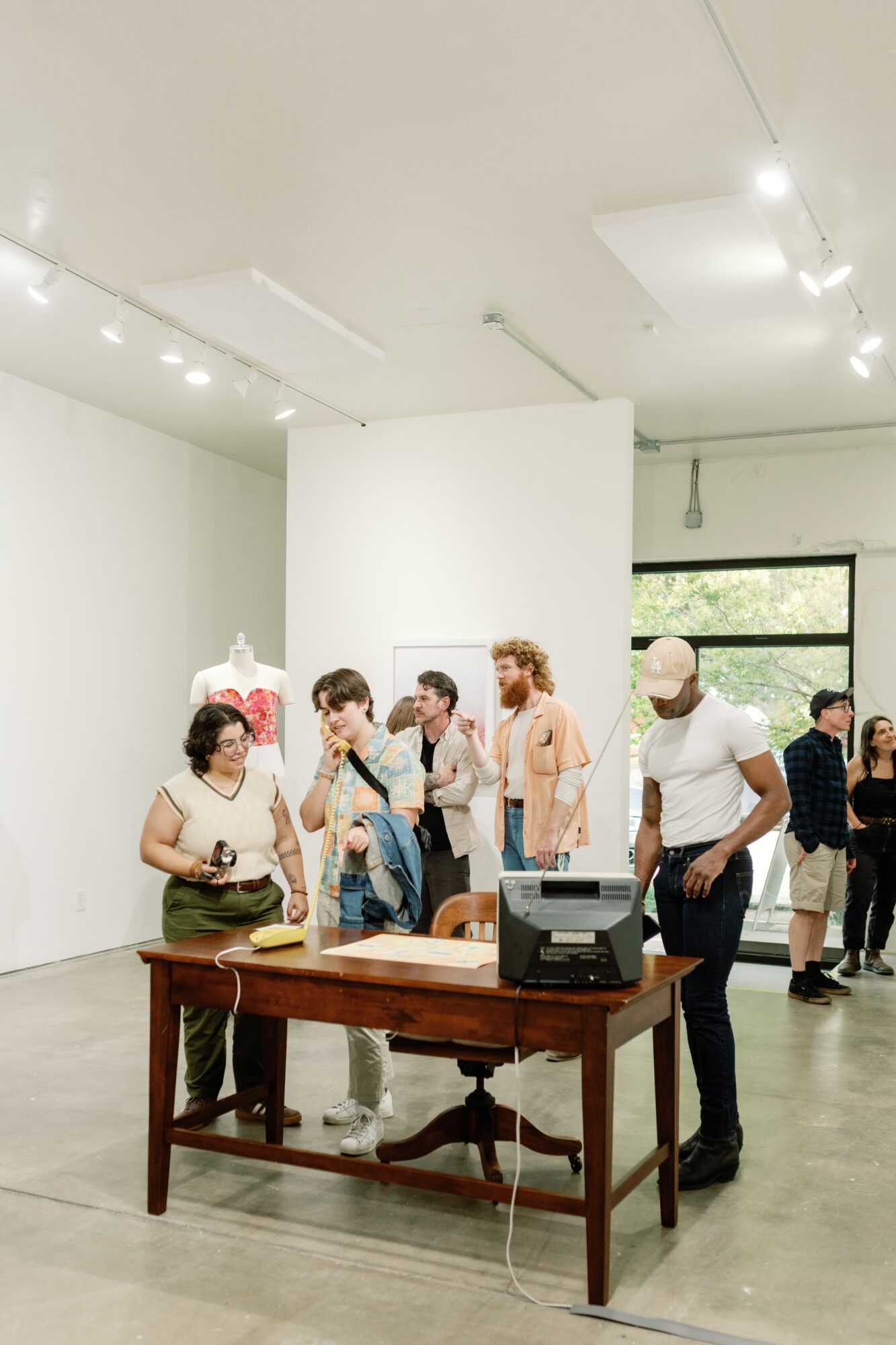Group of people in an art gallery, some looking at artwork, with a wooden table and a cash register in the foreground.