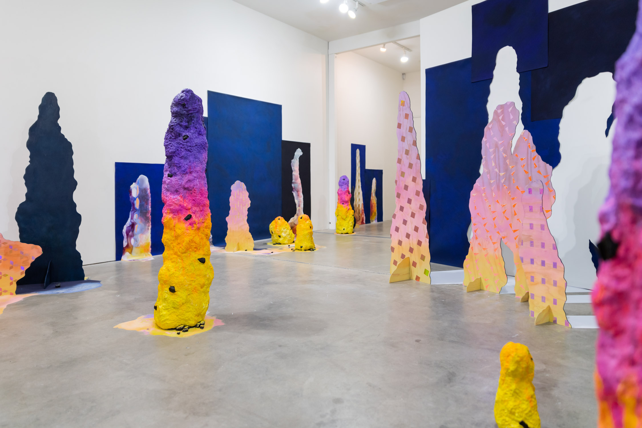 Gallery with colorful, textured sculptures and blue panels, concrete floor, white walls, and ceiling lights.