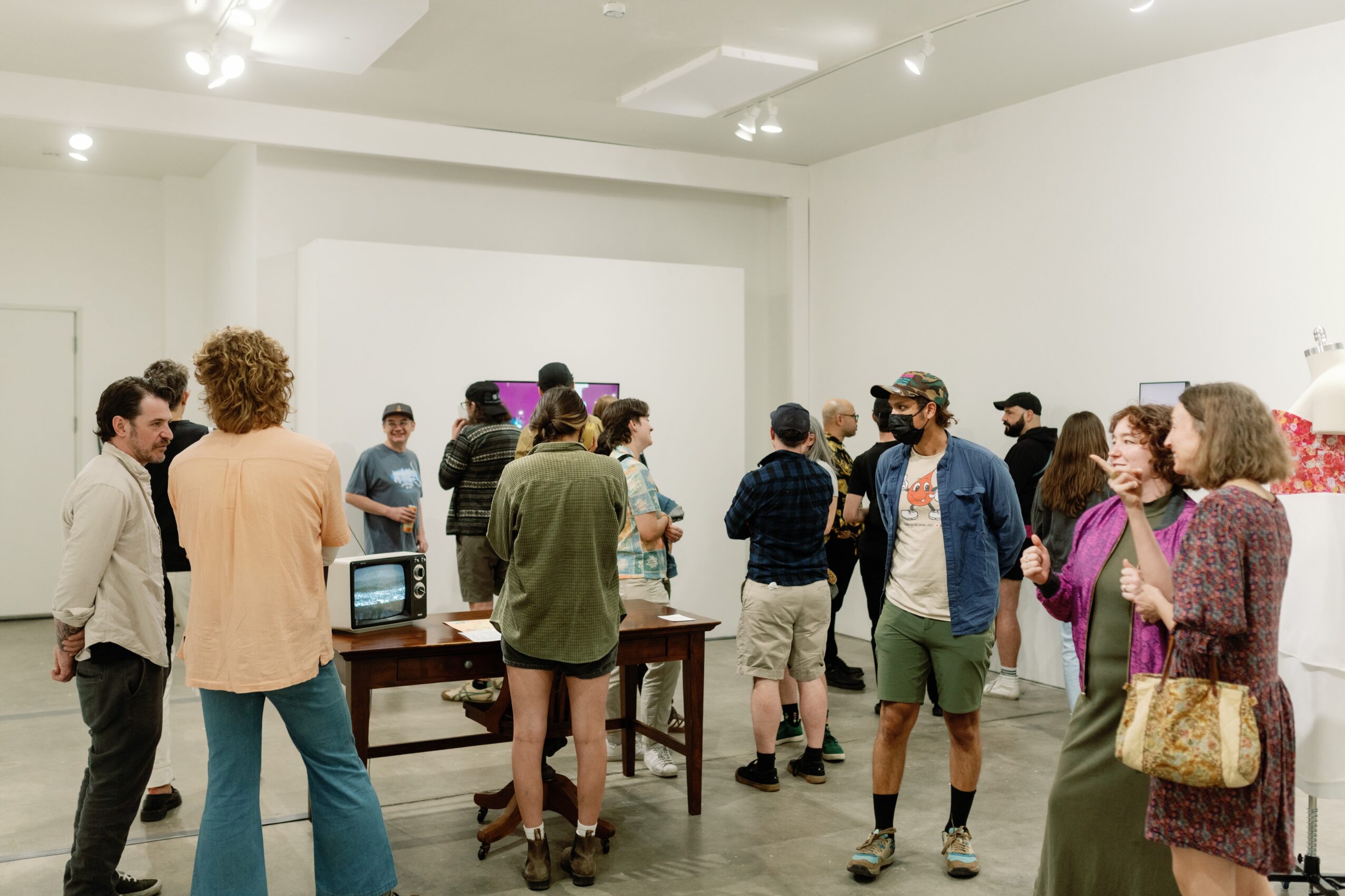 Group of people gathered in a white-walled room, some talking and others observing displays or objects on tables.