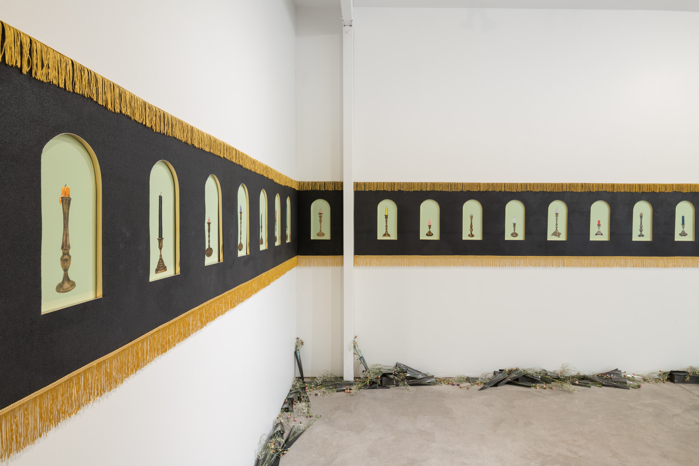 Gallery wall with small framed pictures and candles, black background with gold fringe, white walls, and a damaged floor area.
