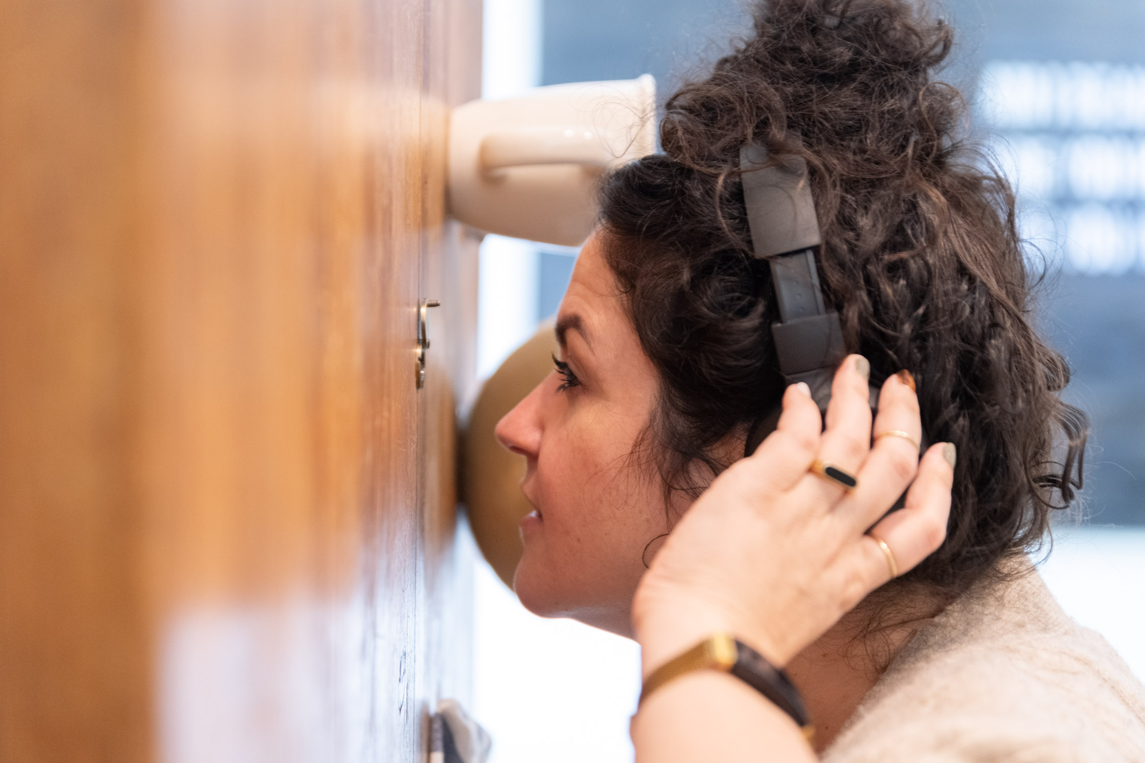 Woman with curly hair wearing headphones, close to a wall, listening intently, profile view, indoor setting.