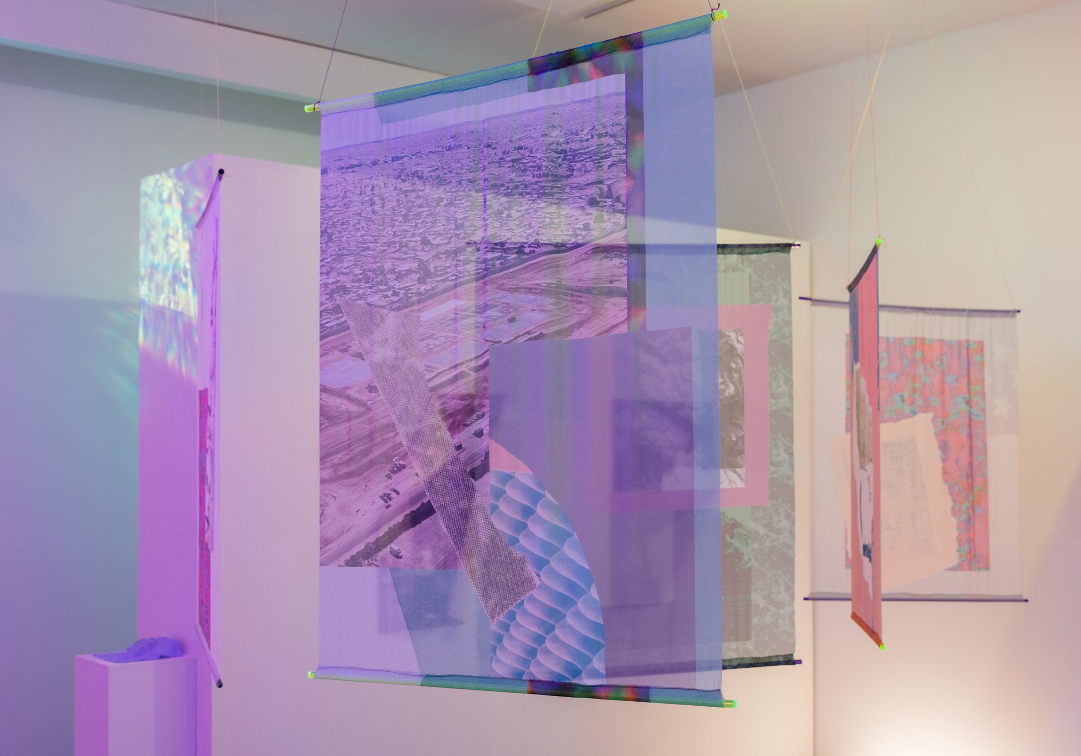 Multiple colorful hanging fabric panels in an art gallery, with a person in a striped shirt reflected in the central panel.