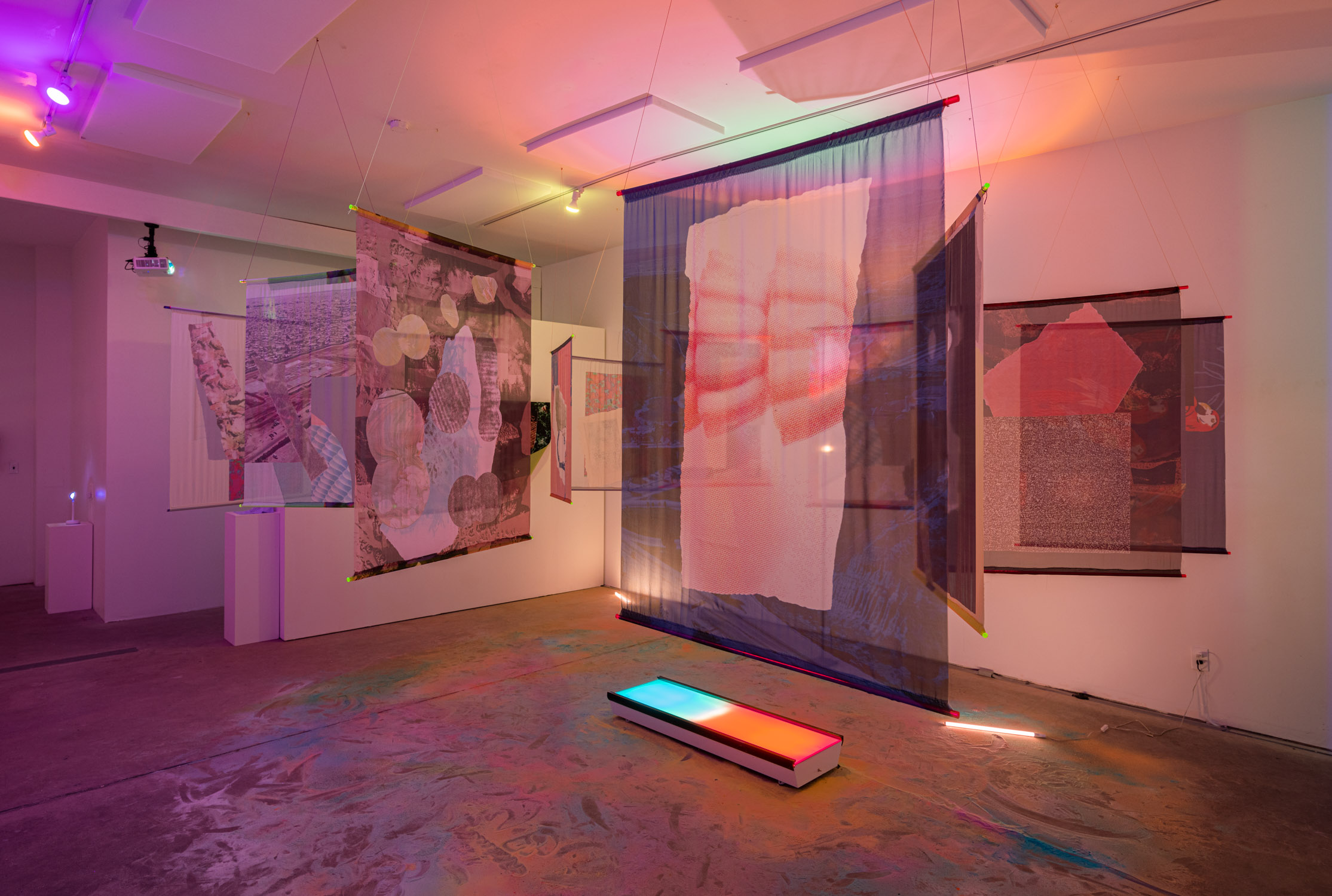 Art installation with hanging fabric and framed artworks in a gallery, illuminated by colorful lighting.