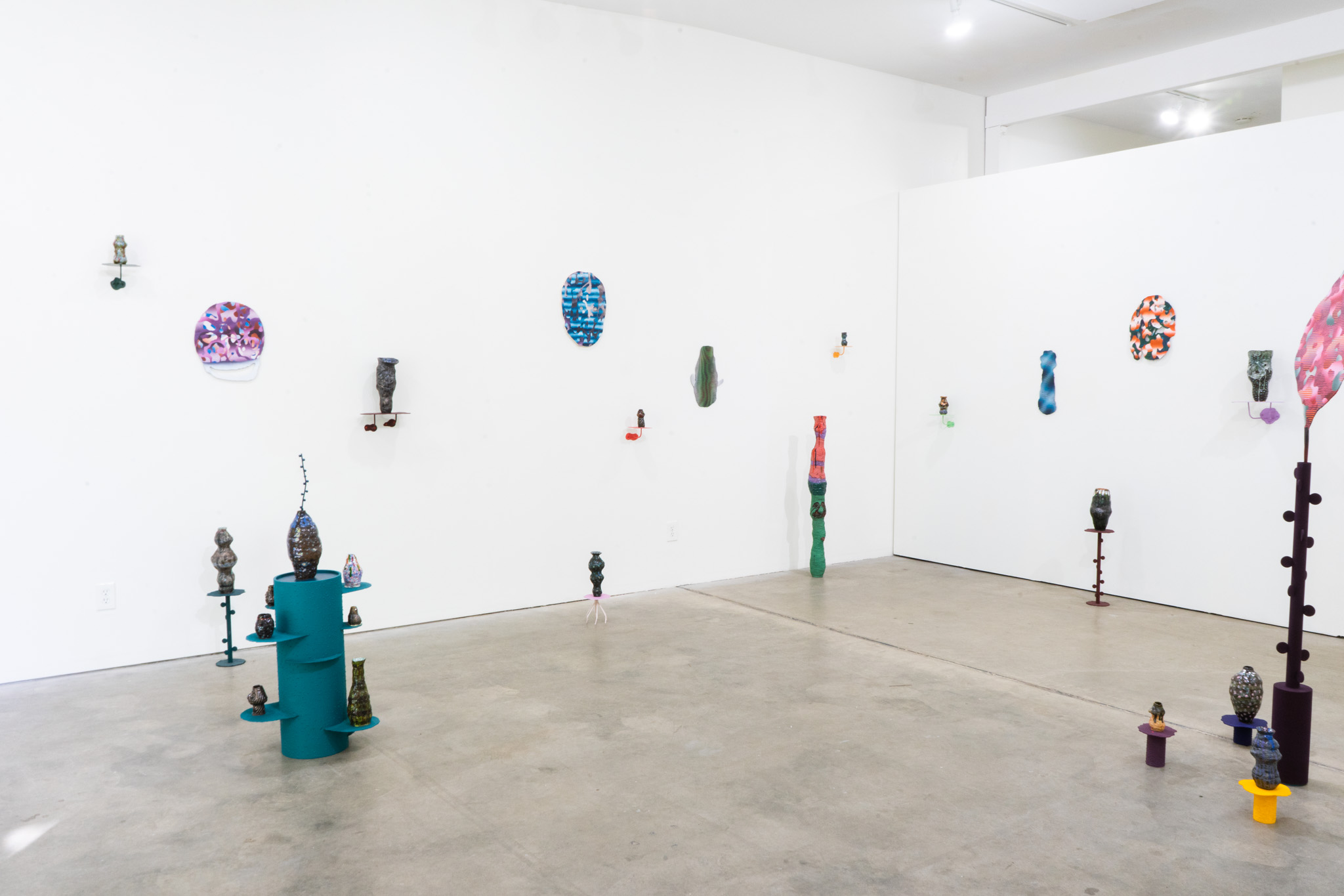 Gallery with white walls displaying colorful abstract sculptures on pedestals and wall mounts, with a concrete floor and bright lighting.