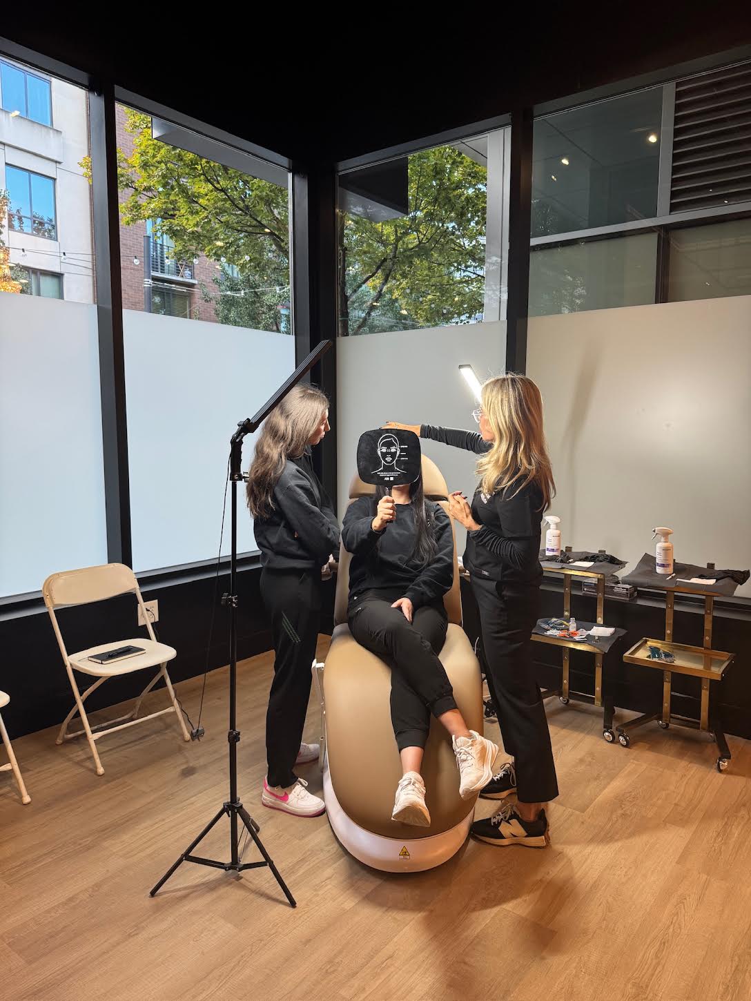 Three women in a room with large windows, one sitting on a chair, two standing, with makeup tools and chairs nearby.